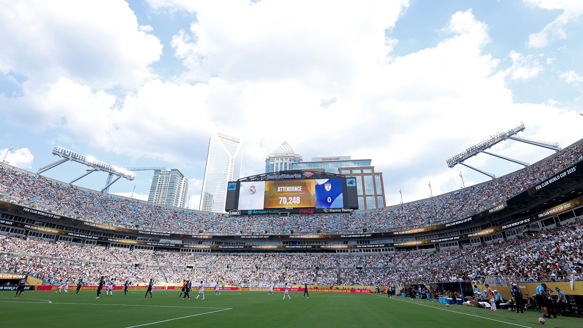 A packed soccer stadium during a match with a scoreboard showing 70,248 attendance and score 0-0 under blue sky with white clouds and city skyscrapers in the background.