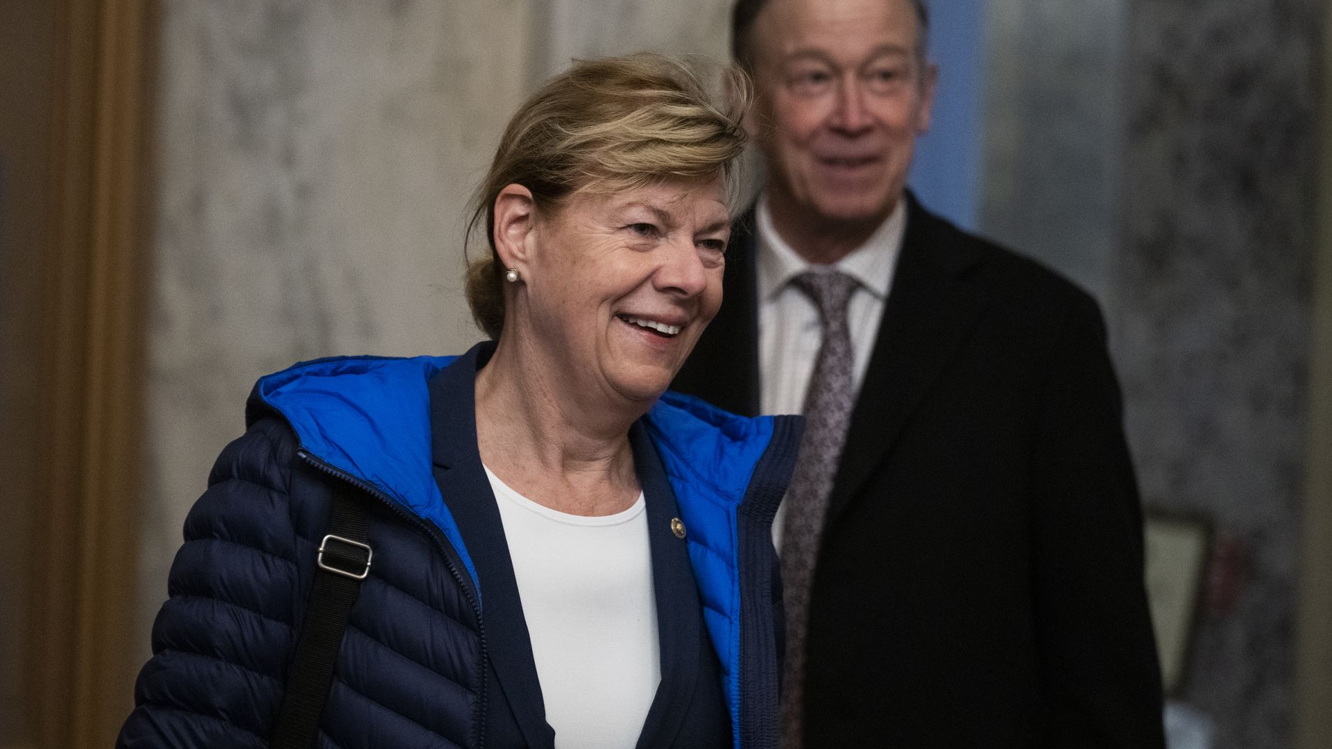 Sen. Tammy Baldwin, wearing a black puffy jacket with a blue lining over a dark blue blazer and white shirt, walks into the capitol in front of Sen. John Hickenlooper.