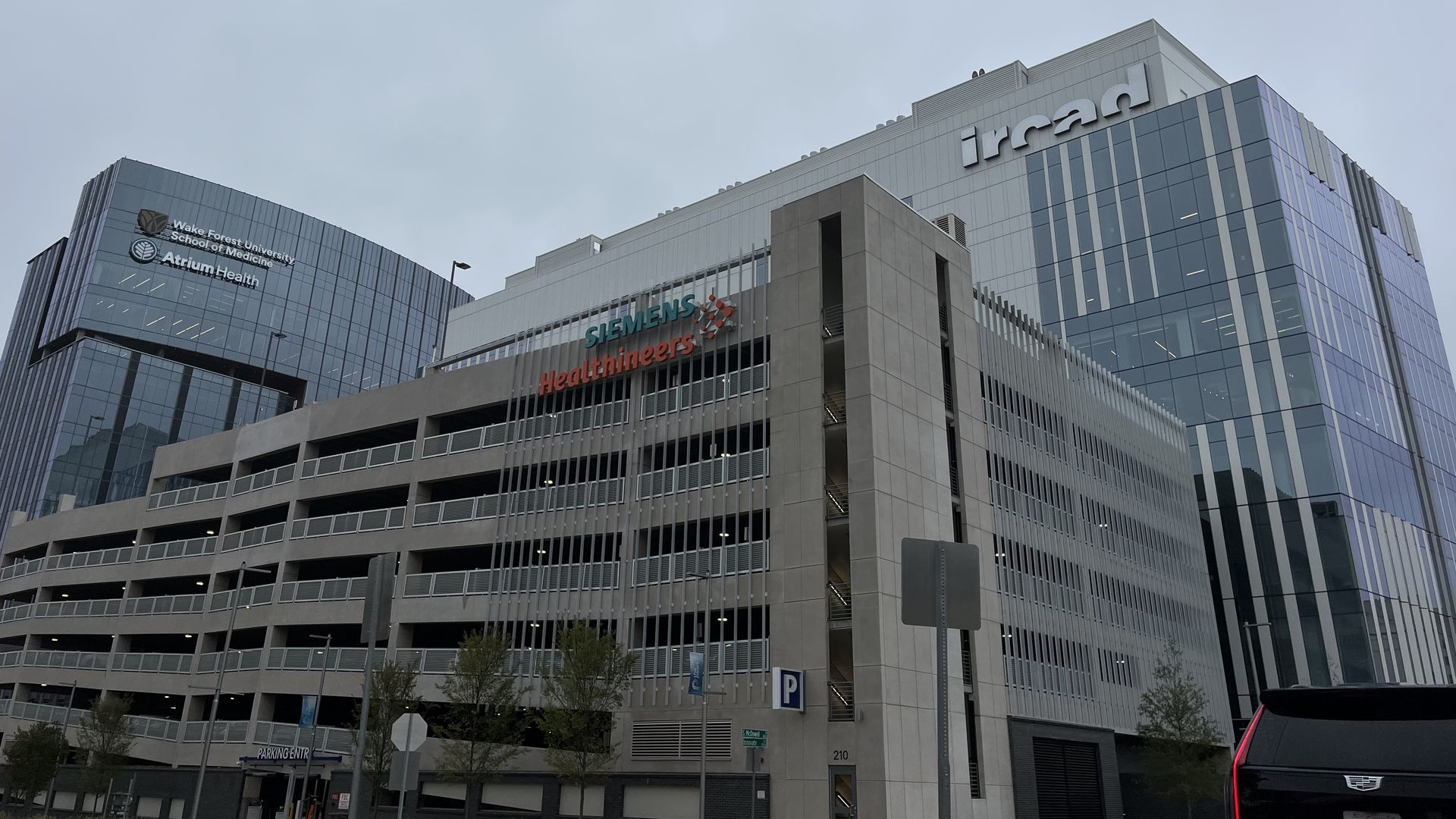 Modern multi-story buildings including Wake Forest University School of Medicine, Atrium Health, Siemens Healthineers, and Ircad with a parking garage and a black vehicle in foreground under gray sky.