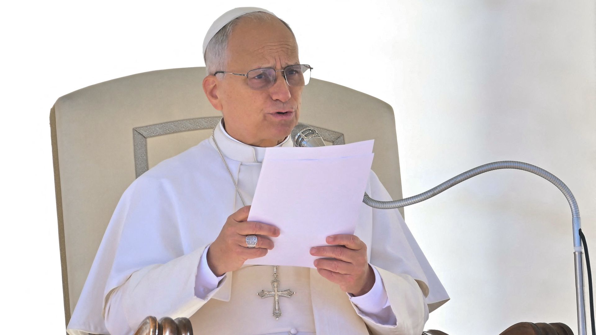 A short, gray-haired, bespectacled Pope Leo XIV, wearing a white cap and gown and large, silver crucifix, speaks into a silver mic while seated on a cream seat with brown, wooden arms.