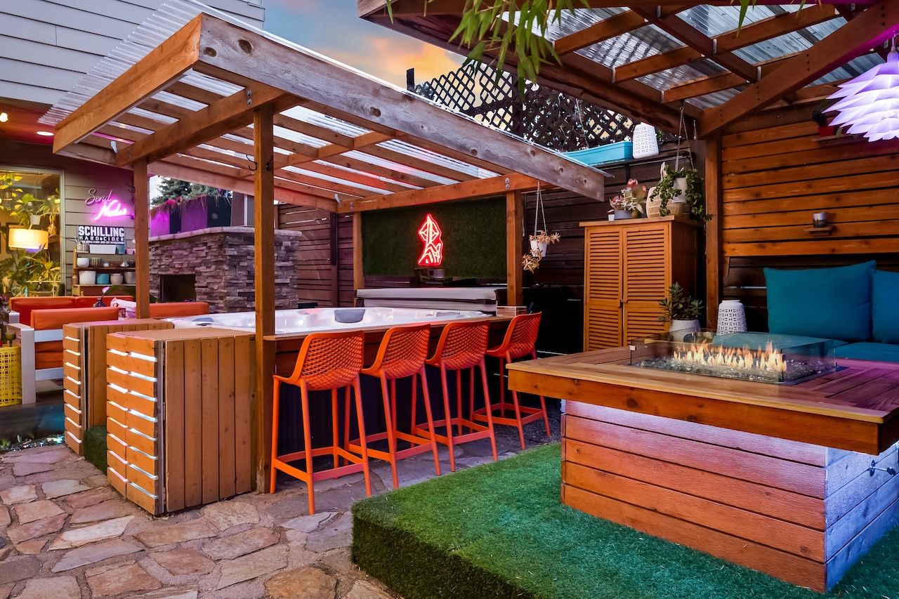 Cozy outdoor patio at sunset with wooden pergola, orange bar stools, a stone floor, a built-in hot tub, and a wooden fire pit table beside blue cushioned seating.