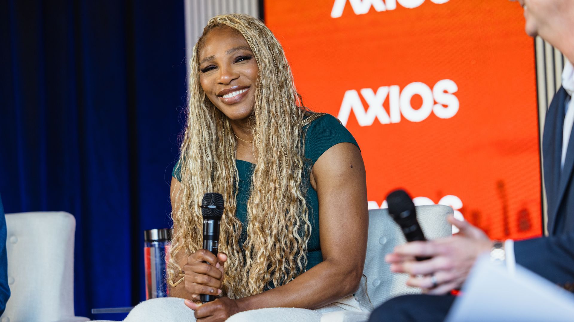 Smiling woman with long blonde dreadlocks in a teal top sits on a stage holding a microphone; a man in a suit sits nearby with another mic, blue curtains and an orange Axios backdrop behind.