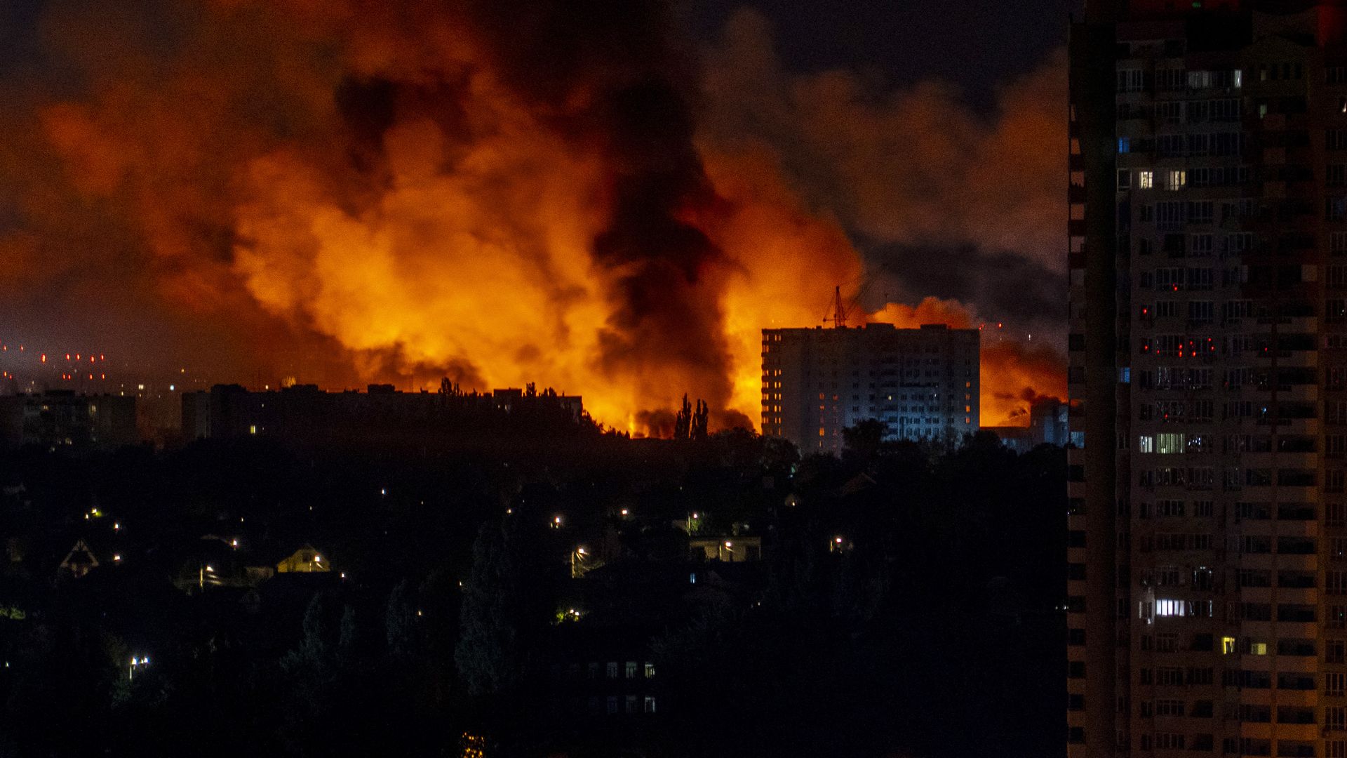 Large fire with thick black smoke billowing behind buildings at night. A cityscape illuminated by orange flames and some lights in surrounding houses.
