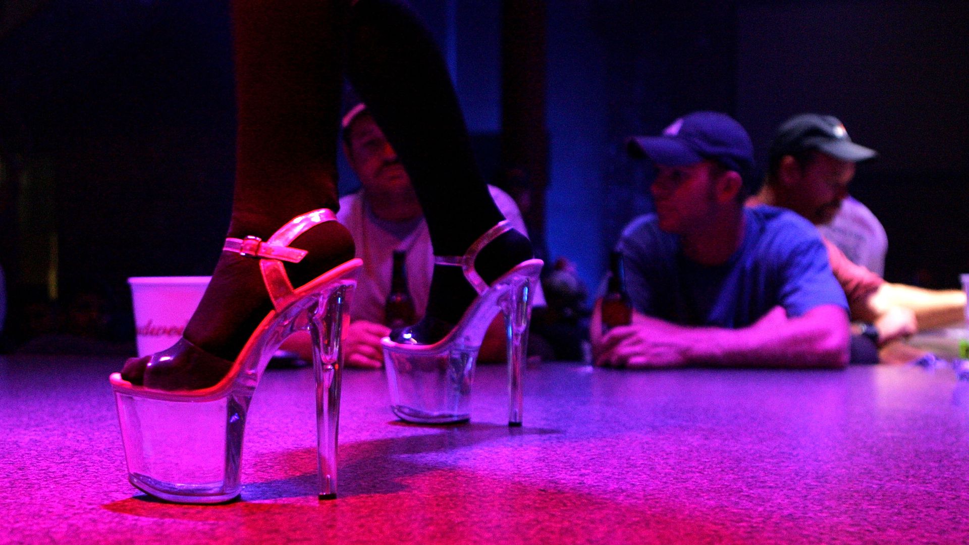 Photo shows a pair of tall, clear platform heels with men sitting behind a bar.