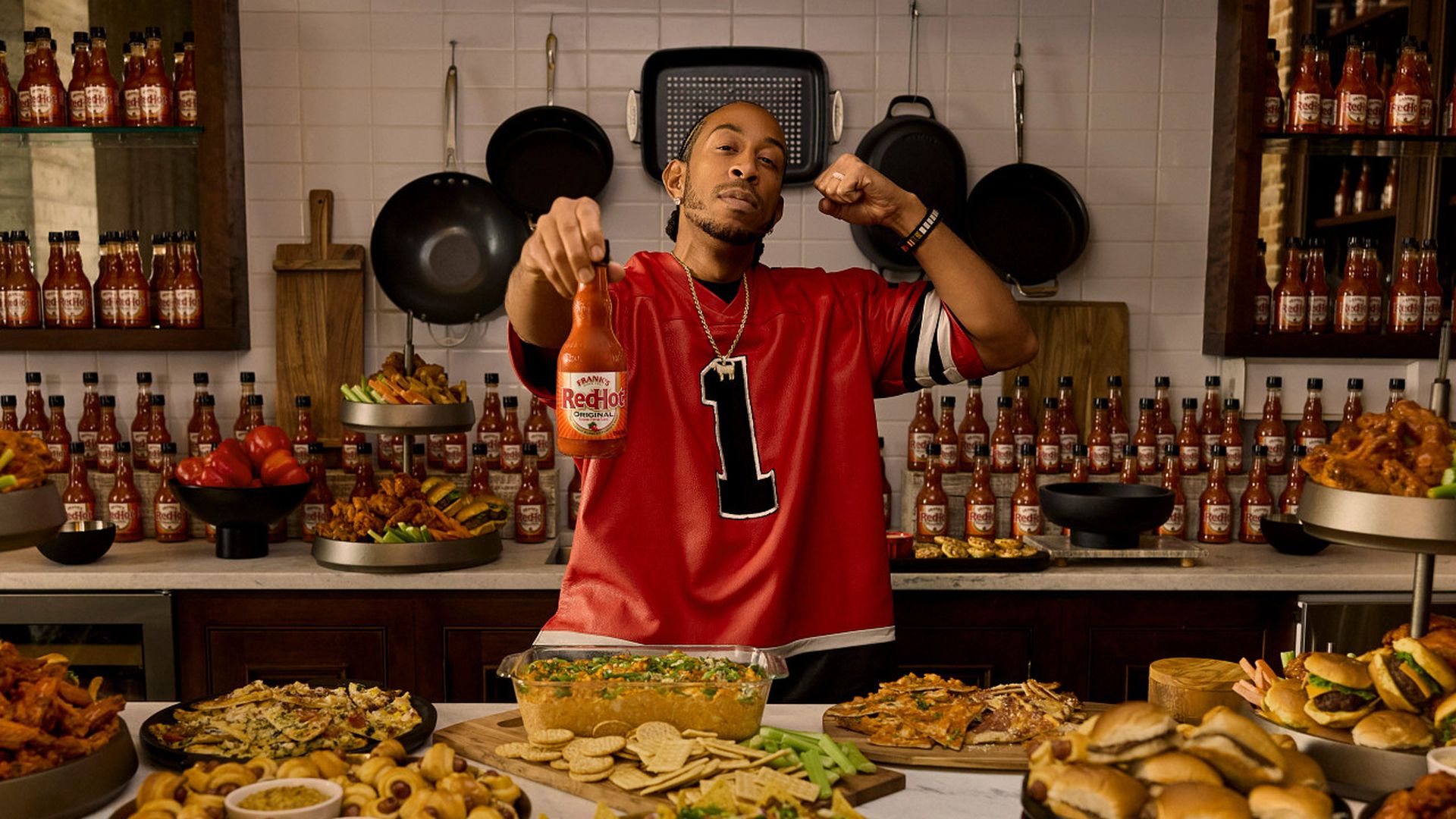 Man in red sports jersey holding a bottle of Frank's RedHot sauce, standing behind a table filled with various foods including wings, sliders, chips, and pizza, with many hot sauce bottles displayed behind him.