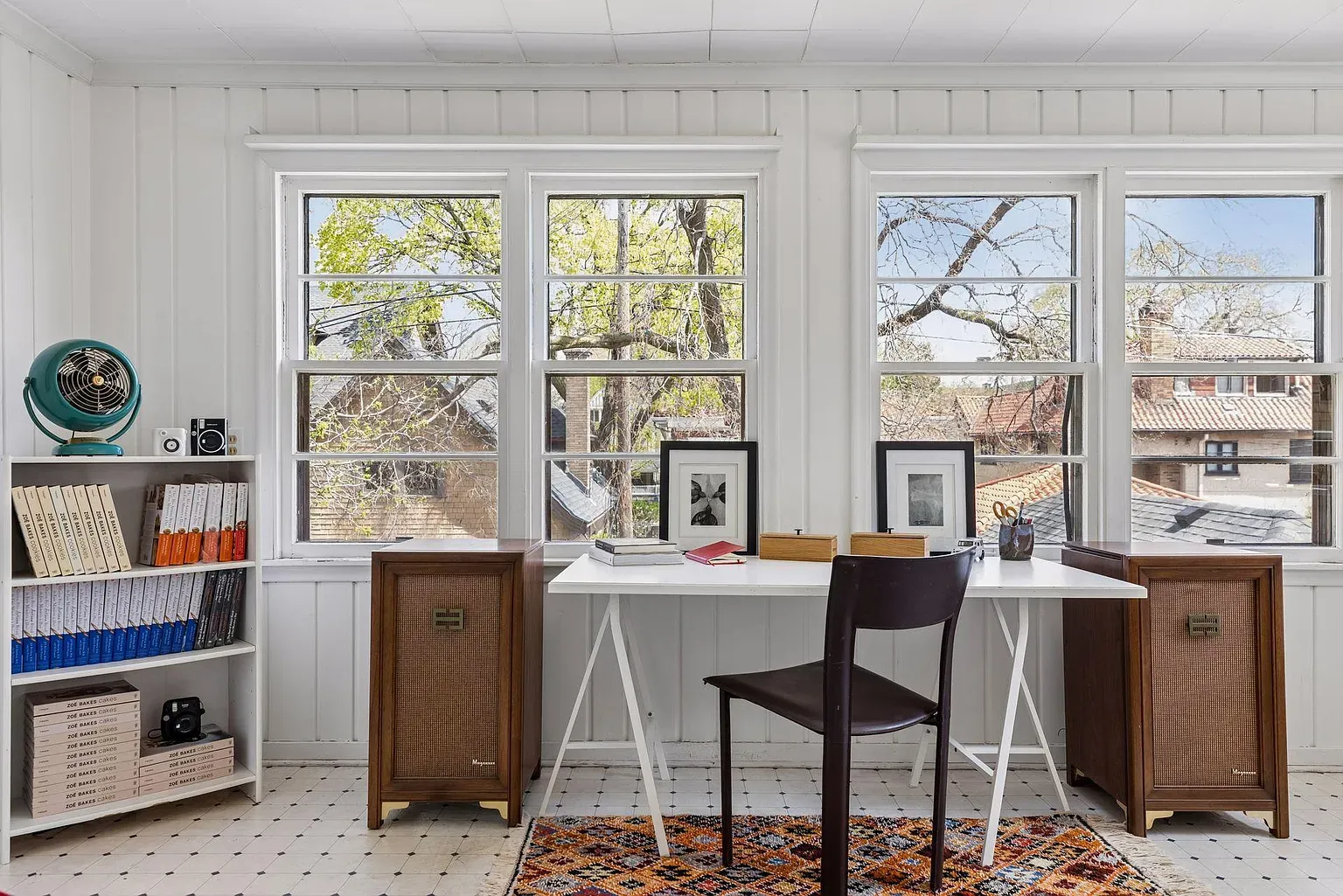 Bright home office with a white desk and black chair centered before three large windows. Vintage cabinets flank the desk, a teal fan on a shelf, shelves of books, framed art, and a patterned rug.