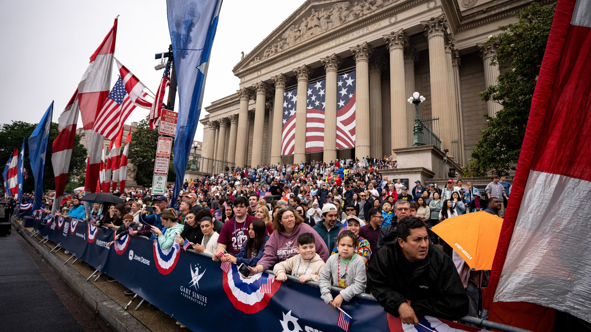 Crowd lines a barricaded street before a grand neoclassical building with tall columns, as large American flags and patriotic banners hang between the columns during a parade.