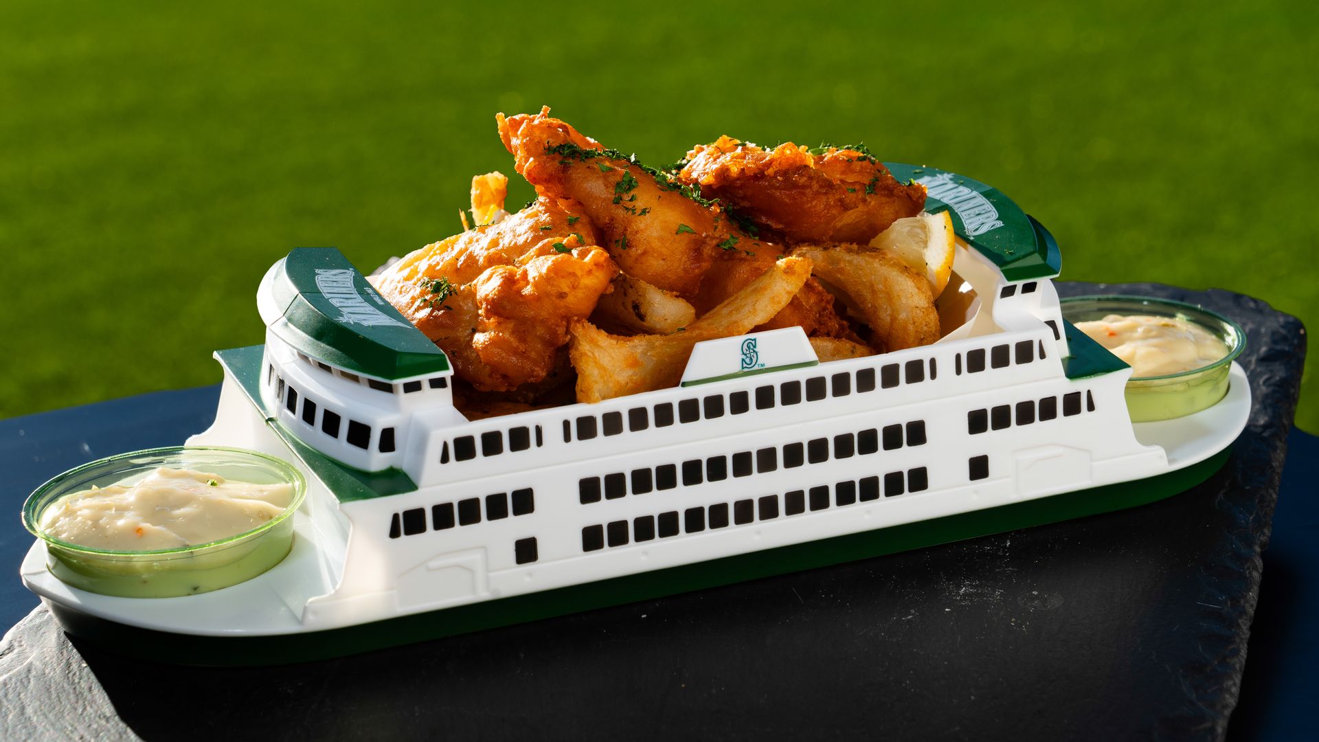 White miniature ferry food carrier with green accents, piled high with fried chicken and potato wedges on its deck, flanked by dipping sauce cups, set on a dark surface with green grass in the background.