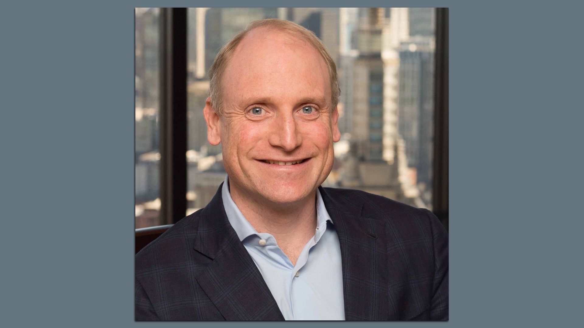 Portrait of a smiling, balding man in a dark blazer and light blue shirt, with a city skyline visible through a window behind him.