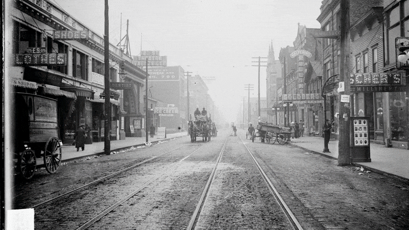 Black-and-white photo of a cobblestone street with horse-drawn carriages, streetcar tracks, pedestrians, and old storefronts including a dentist and milliner in a foggy urban setting.