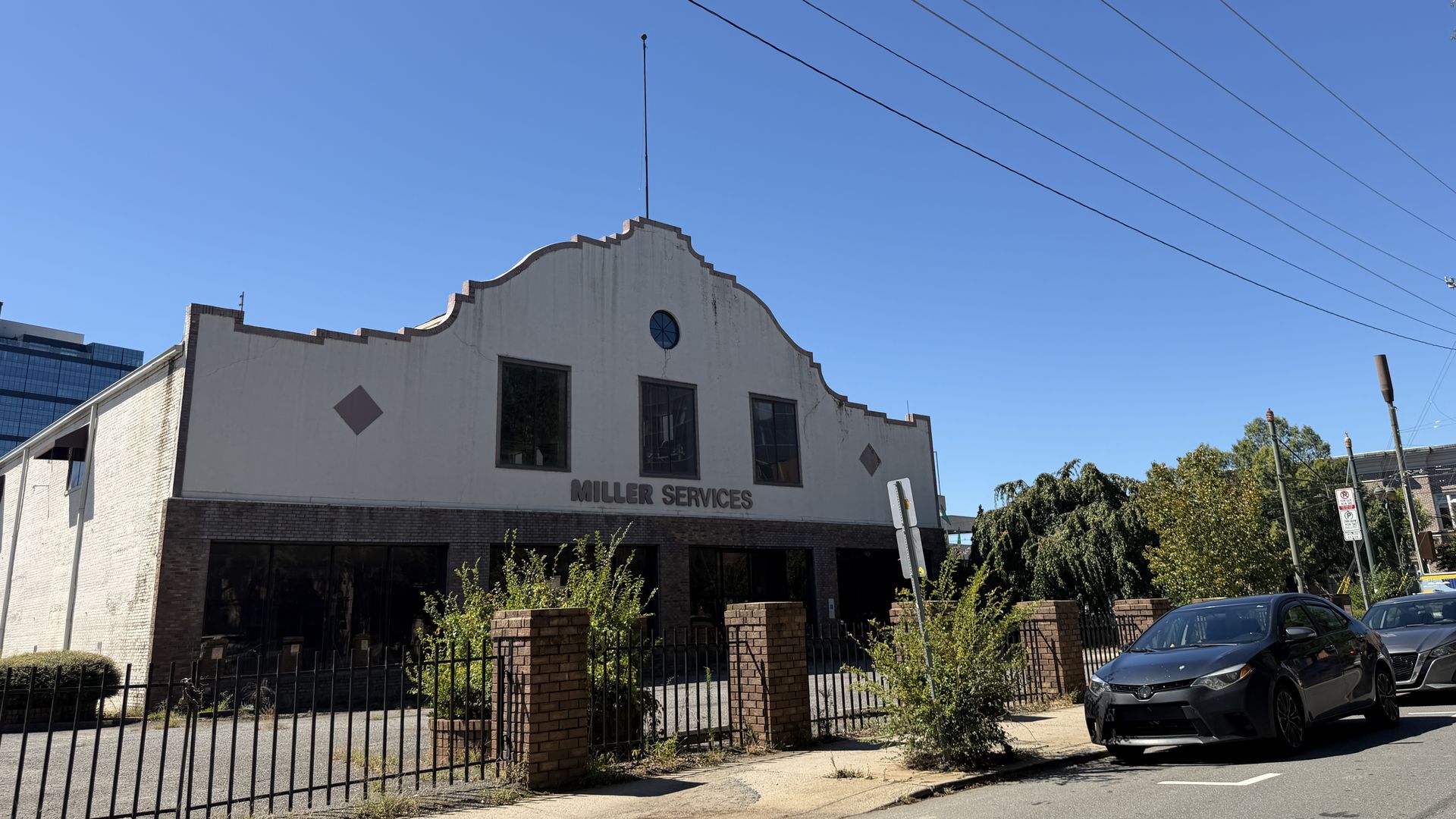 Blue sky over a white, two-story building with a stepped roofline and a circular window; Miller Services on the front, brick base, black fence, and a parked car below power lines.