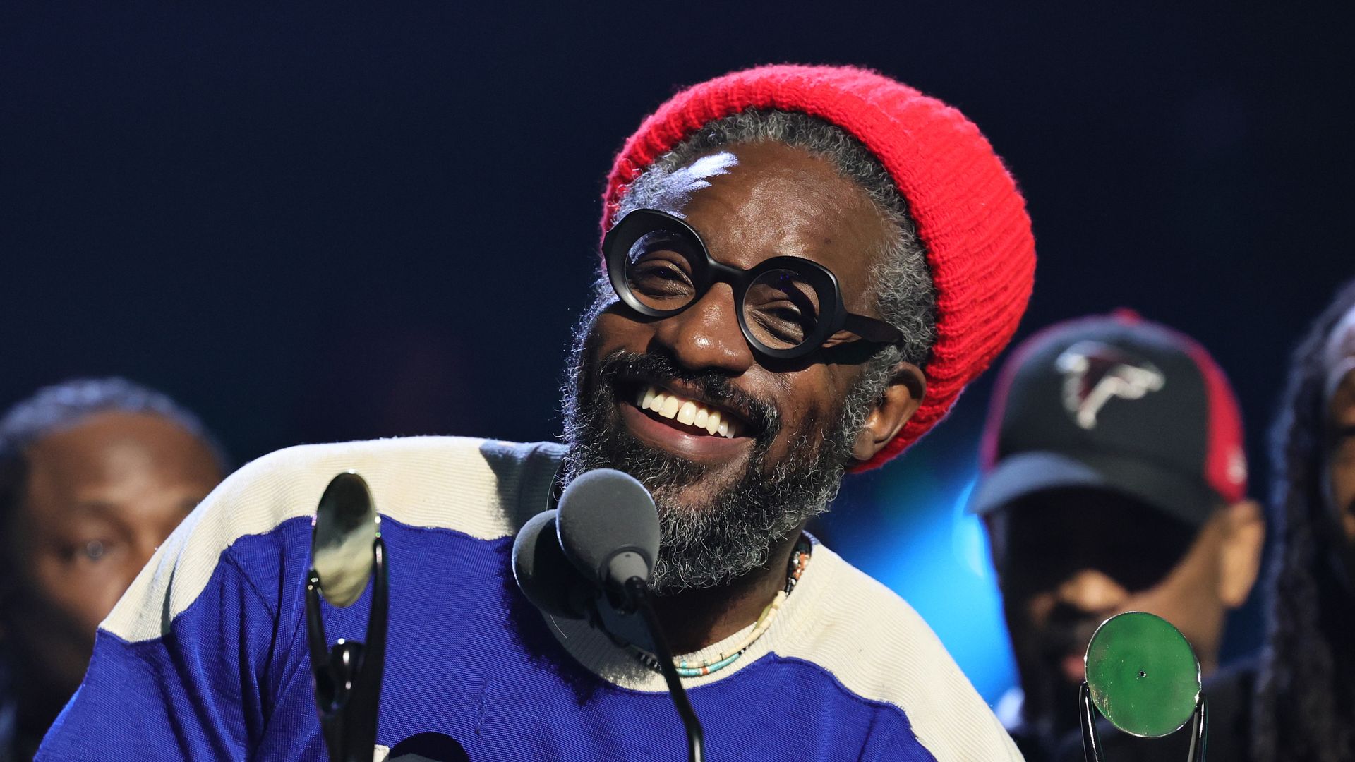 A smiling Black man with salt and pepper hair and beard wearing a red beanie and a blue jersey with white shoulder features. He is speaking into a round fuzzy microphone and backed by several people, including one wearing an Atlanta Falcons baseball hat.