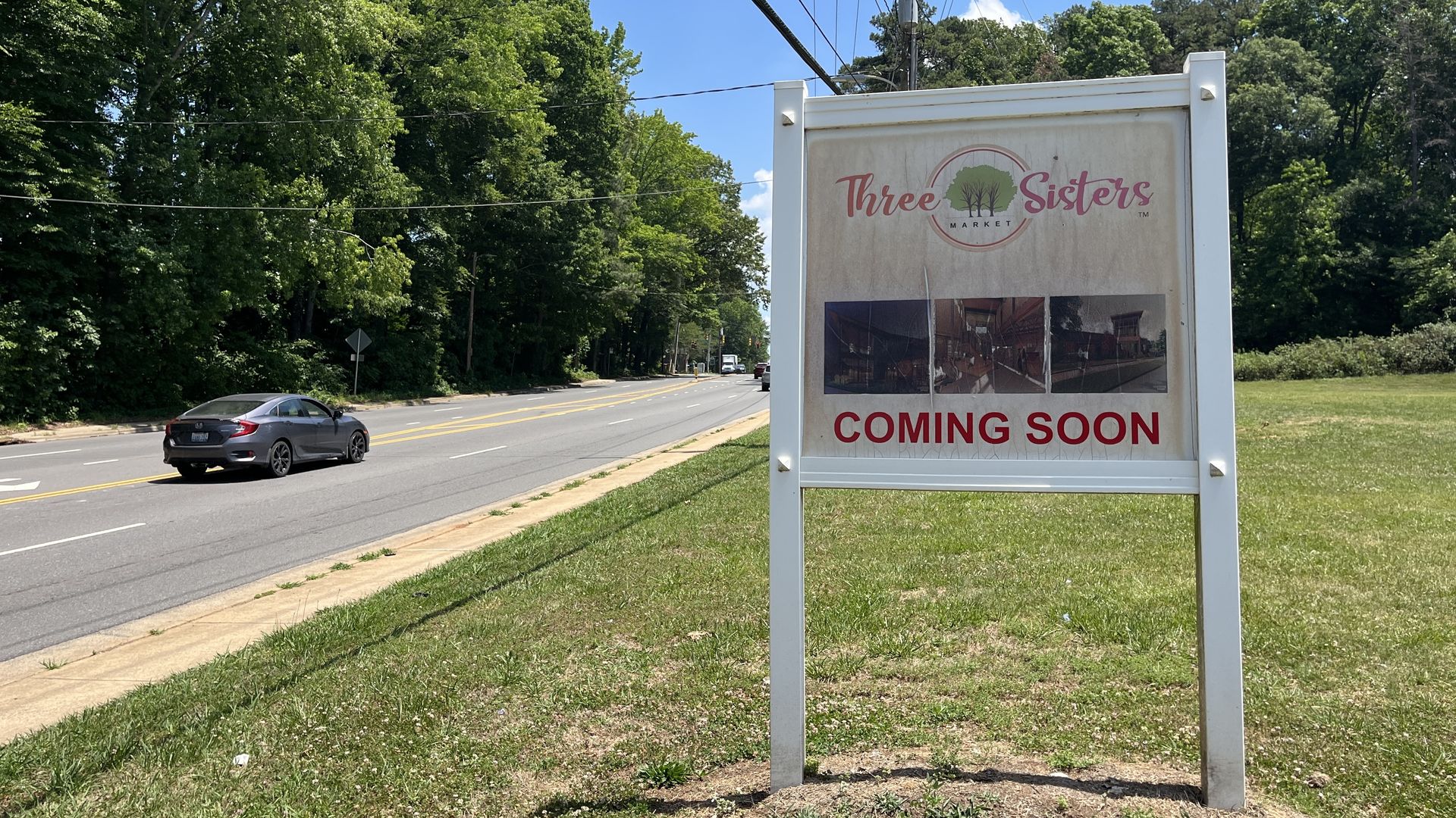 Sunlit street lined with green trees. A large white sign reads "Three Sisters Market" with a tree logo and "COMING SOON" in red; a gray car drives by as power lines cross a blue sky.