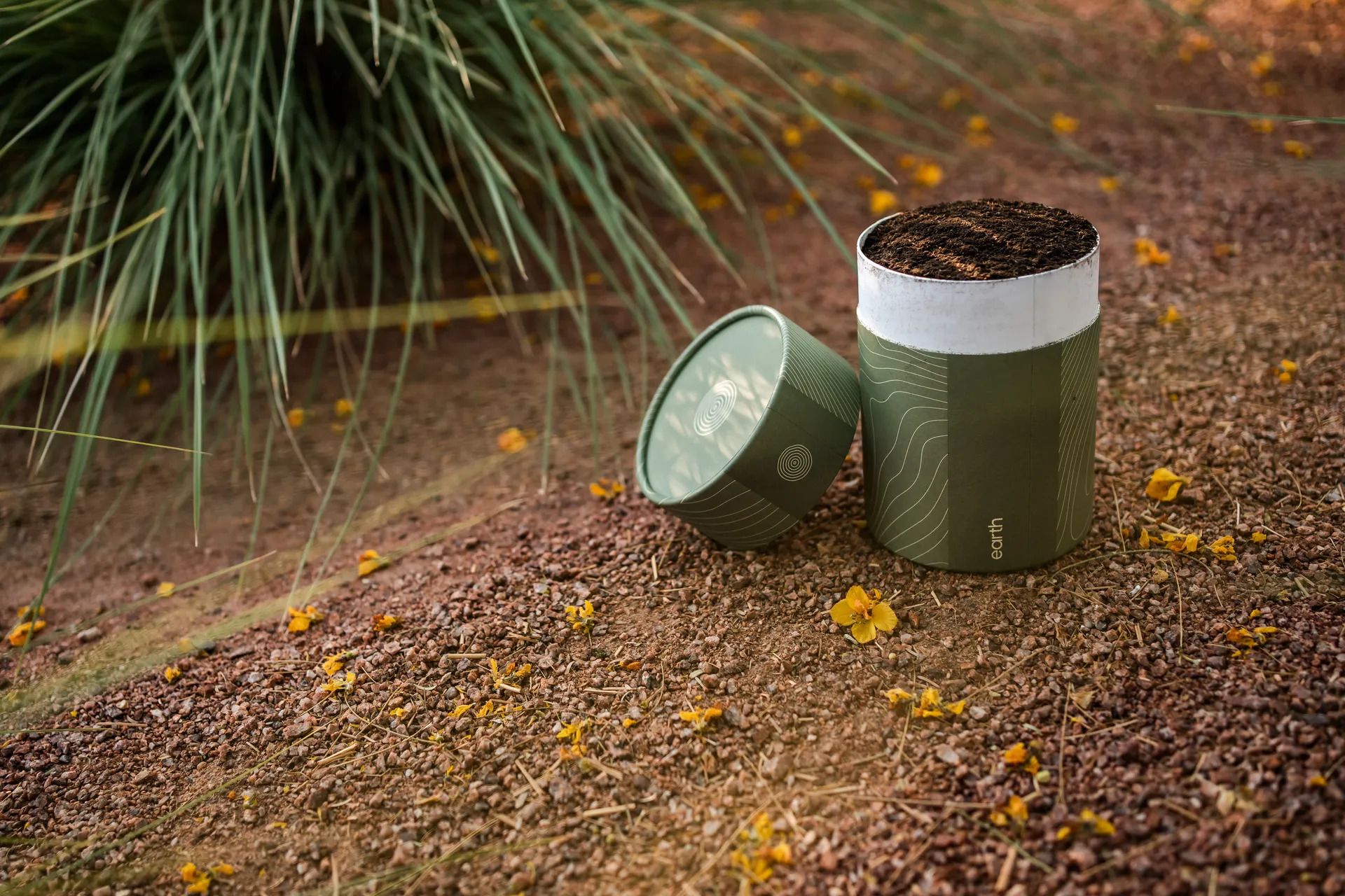 A green cylindrical compost bin on a gravel path with its soil-filled lid removed and leaning next to it; a spiky green plant frames the left, and yellow flowers are scattered on the ground.