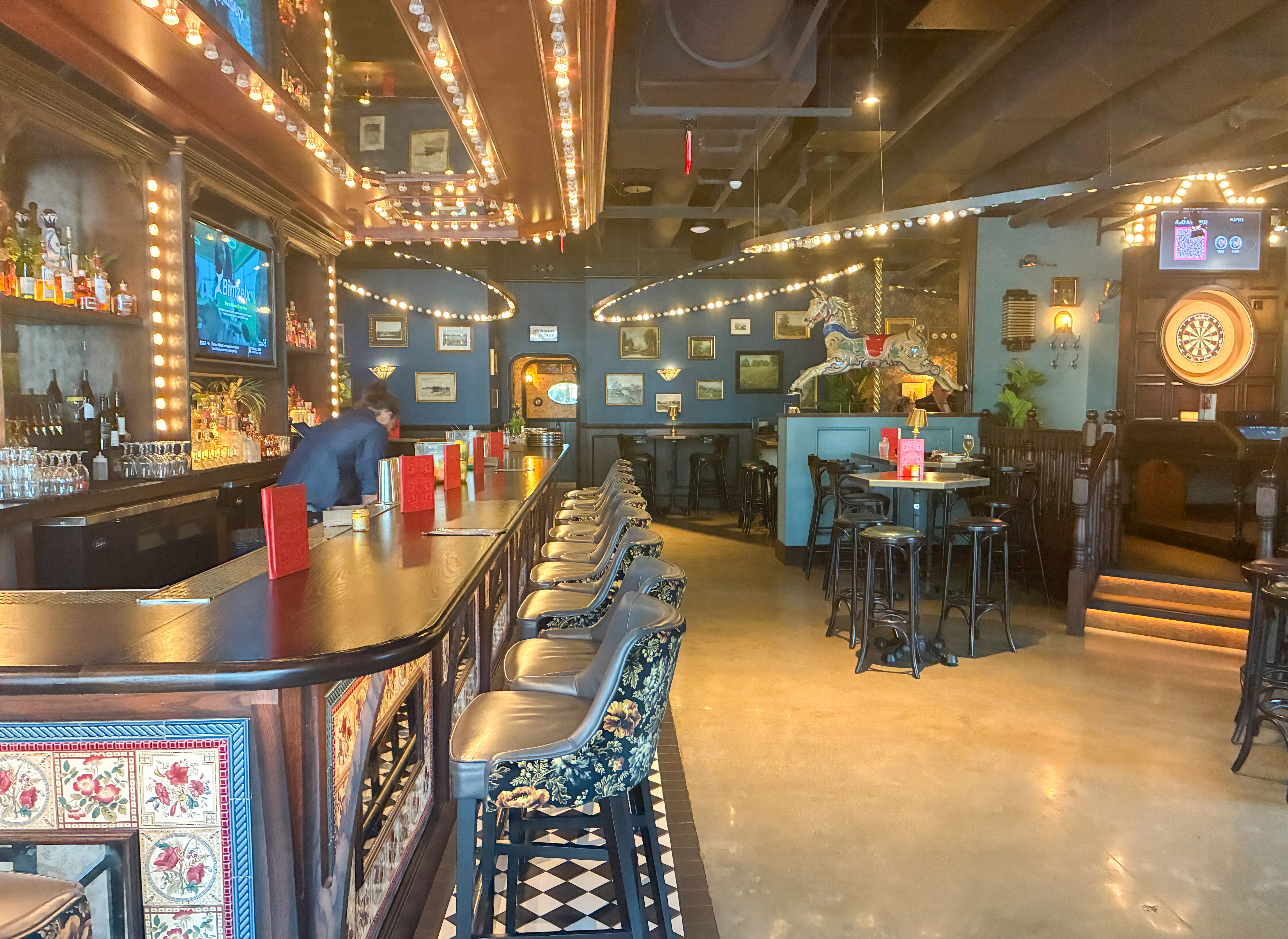 Cozy bar interior with a long wooden counter and metal stools, warm string lights along the ceiling, blue walls lined with framed pictures, a decorative carousel horse, and a dartboard corner.