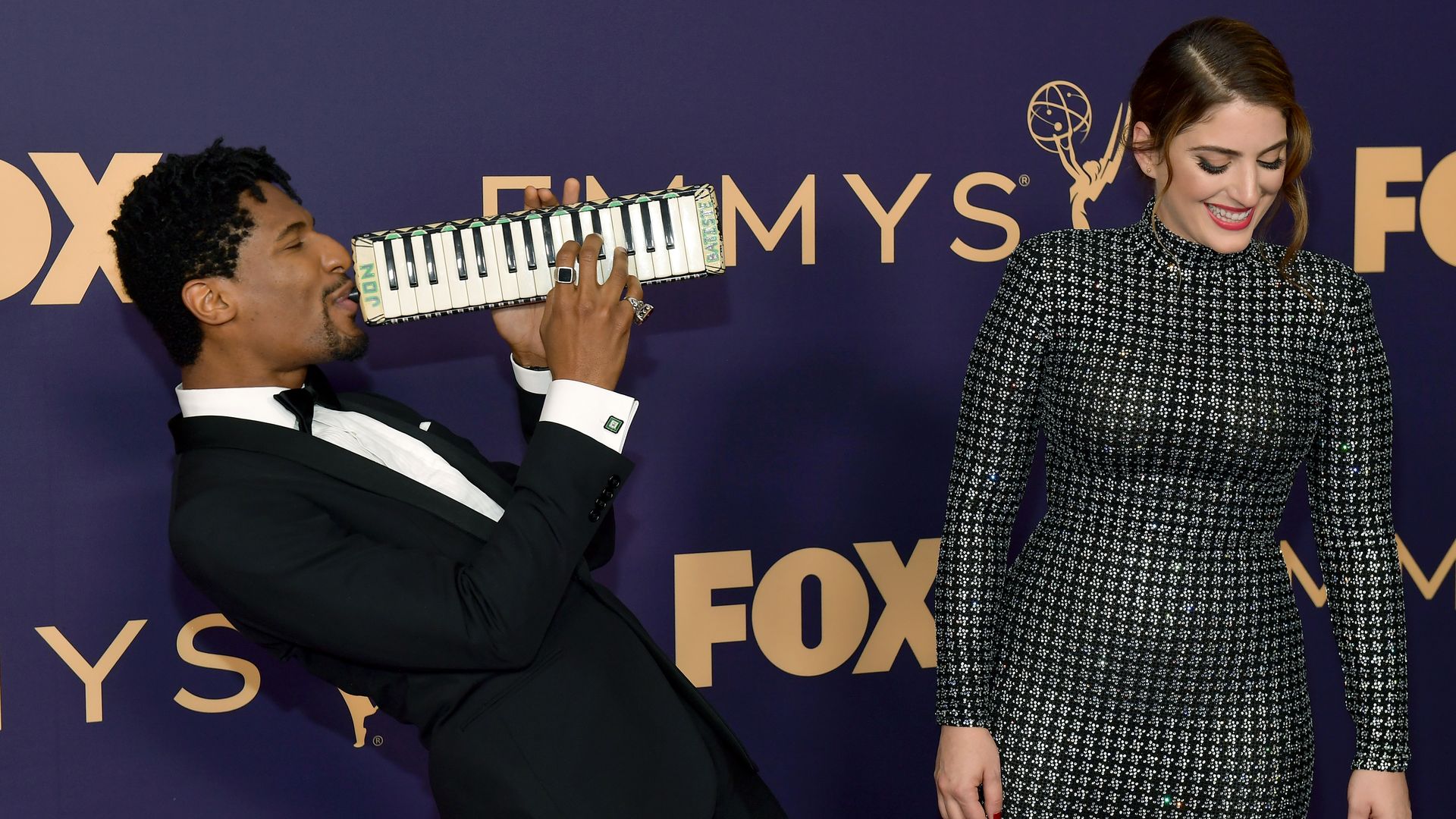 Jon Batiste, wearing a tux, blows into an instrument that looks like a handheld piano, while he walks the red carpet with Suleika Jaouad, who laughs at his antics and looks toward the ground.