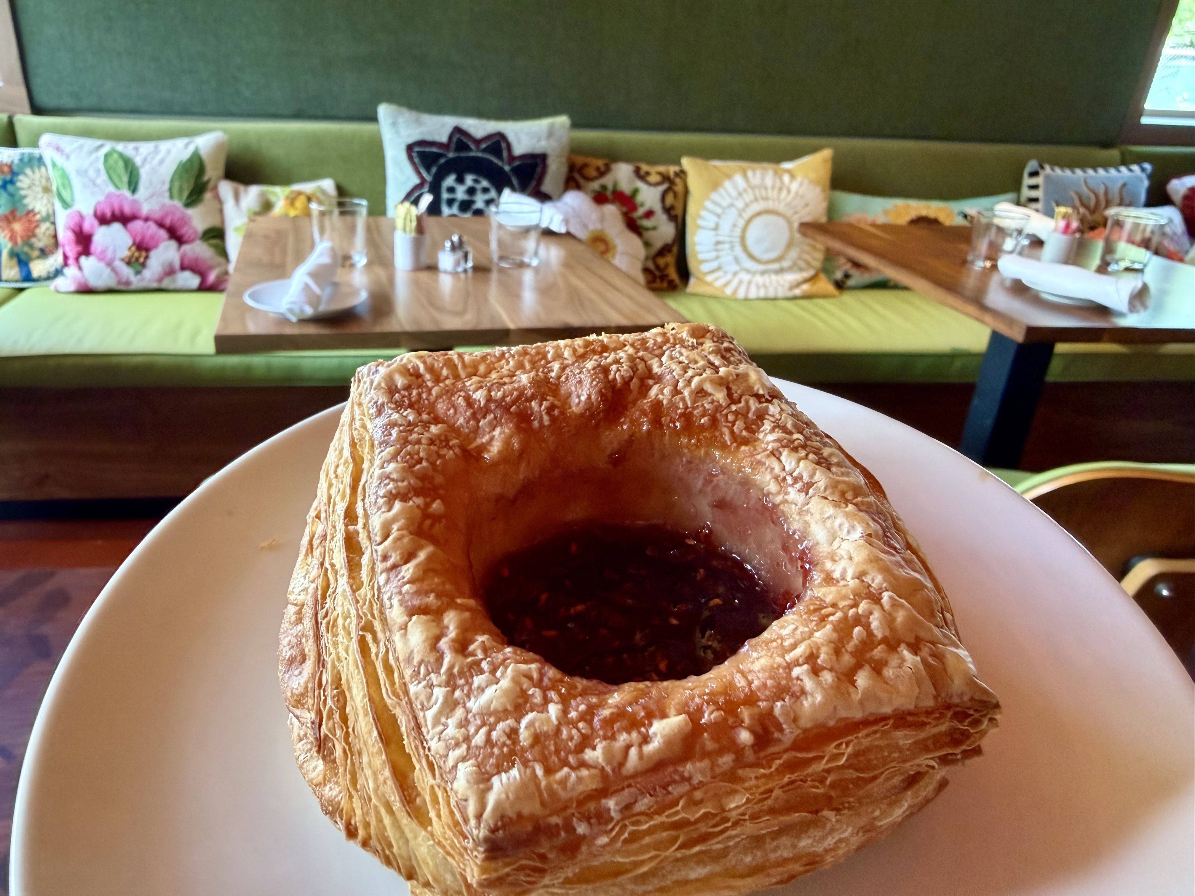 Close-up of a flaky, golden-brown pastry on a white plate in the foreground. In the blurred background, a green couch with colorful patterned cushions and wooden tables in a cozy cafe setting.