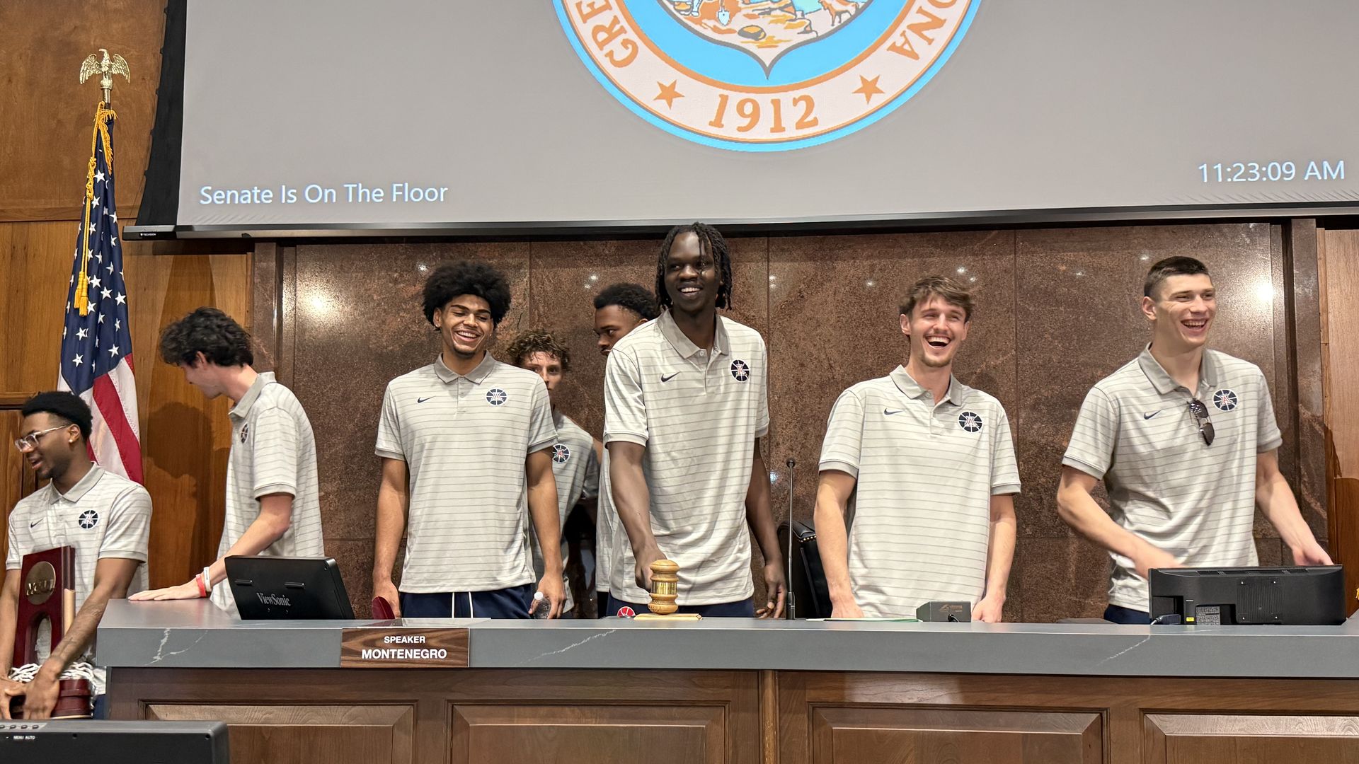 Six young men in gray polo shirts with small University of Arizona logos on them stand smiling behind a gray podium in a wood-paneled legislative chamber; one bangs a gavel. An American flag is to the left; a screen shows a seal and the words "Senate Is On The Floor".