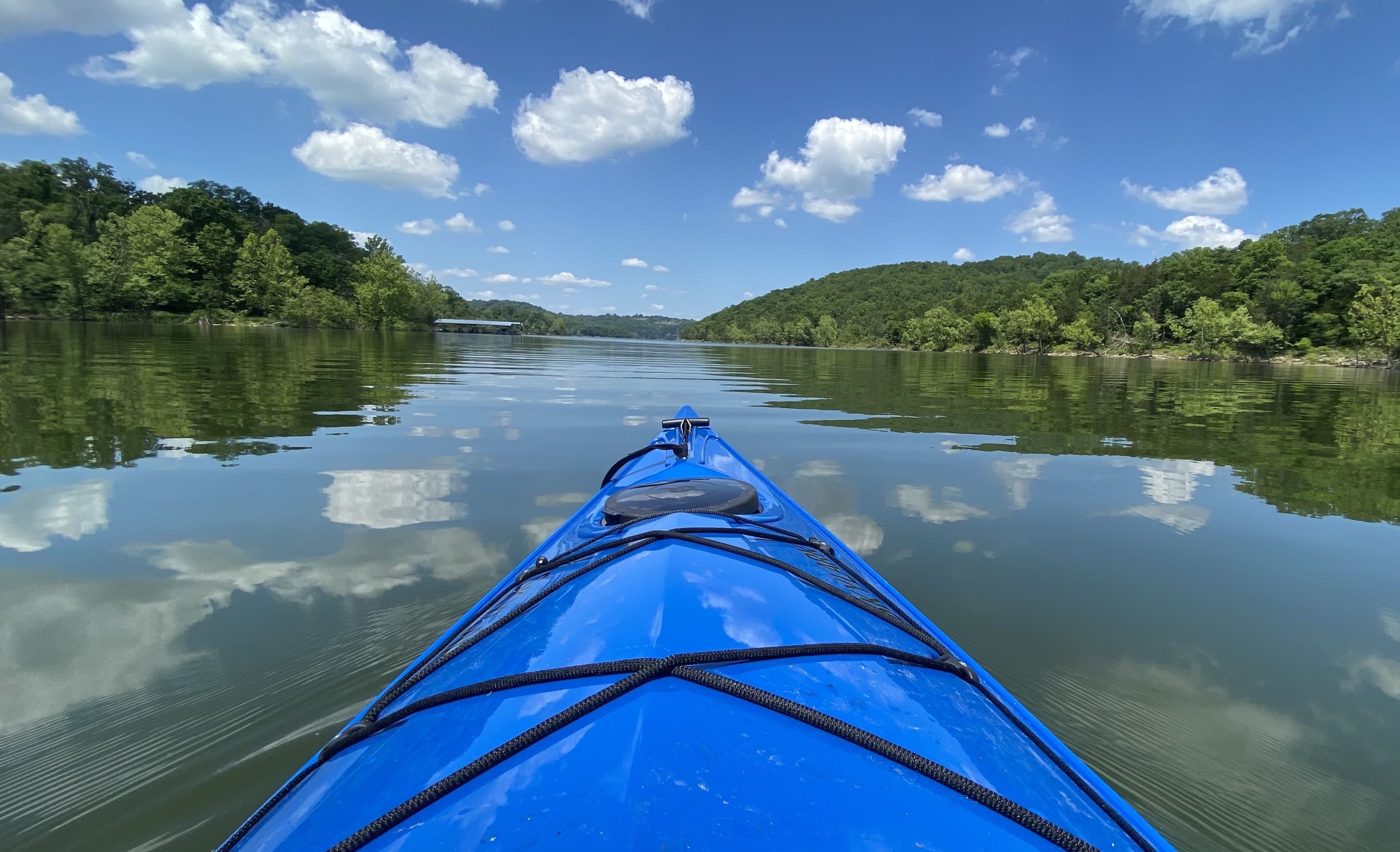 A photo of the front of a blue kayak on water reflecting the clouds in the sky, framed with greenery