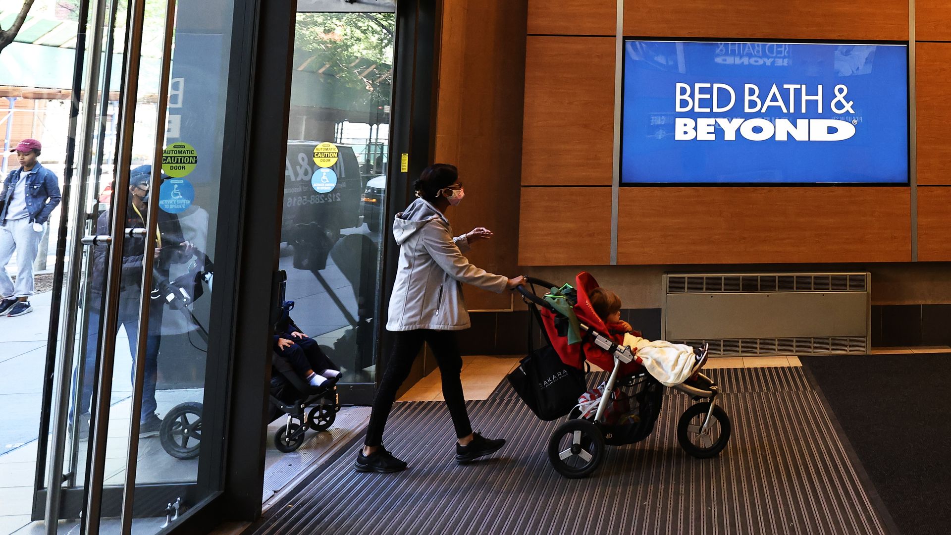 A woman in a tan-colored hooded jacket and black leggings pushes a child in a black baby stroller past a video screen that features the Bed Bath & Beyond name in white lettering on a blue backdrop.
