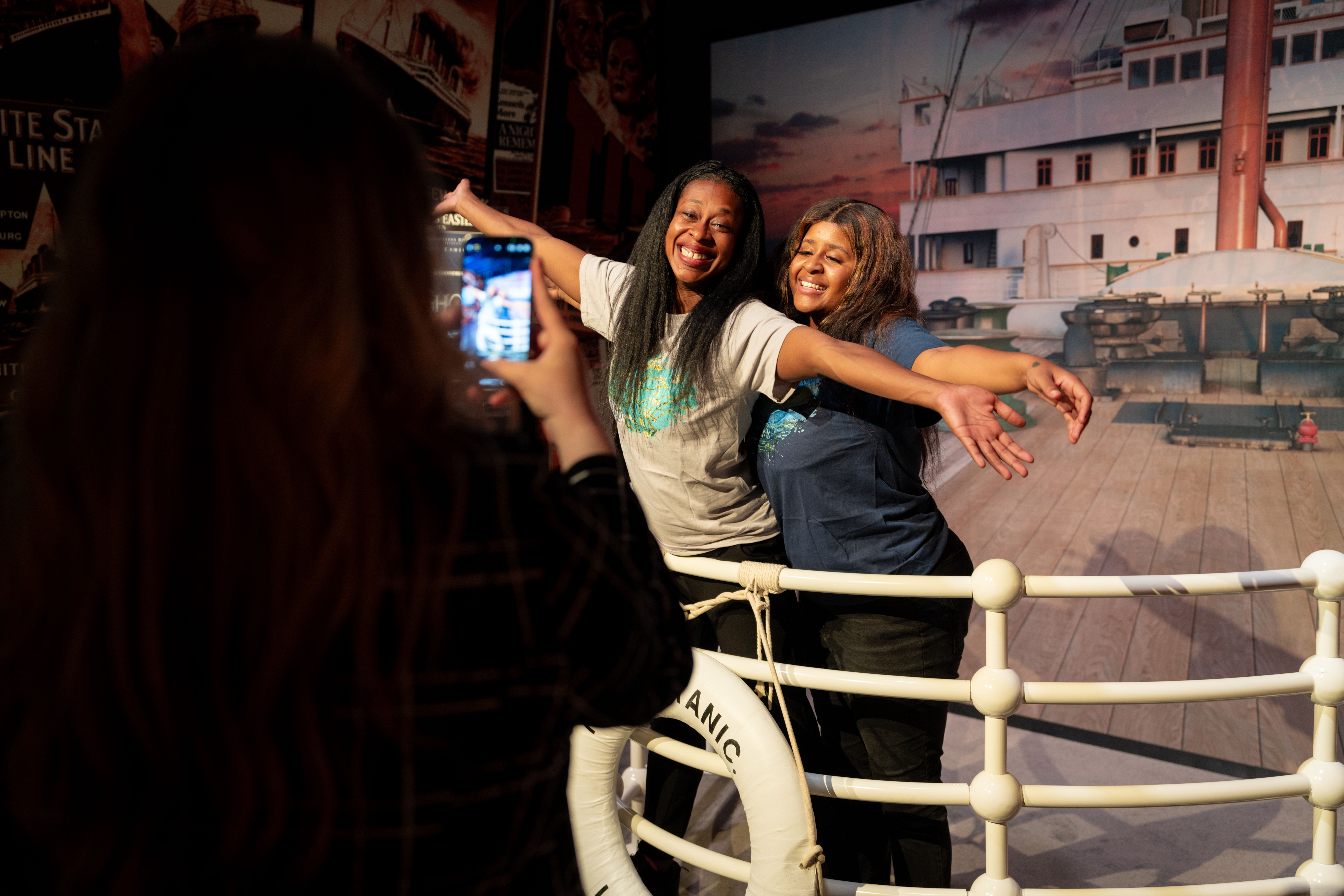 Two smiling women pose with arms outstretched in front of a large ship mural; a person in the foreground uses a smartphone to capture the moment, with a dark silhouette on the left.