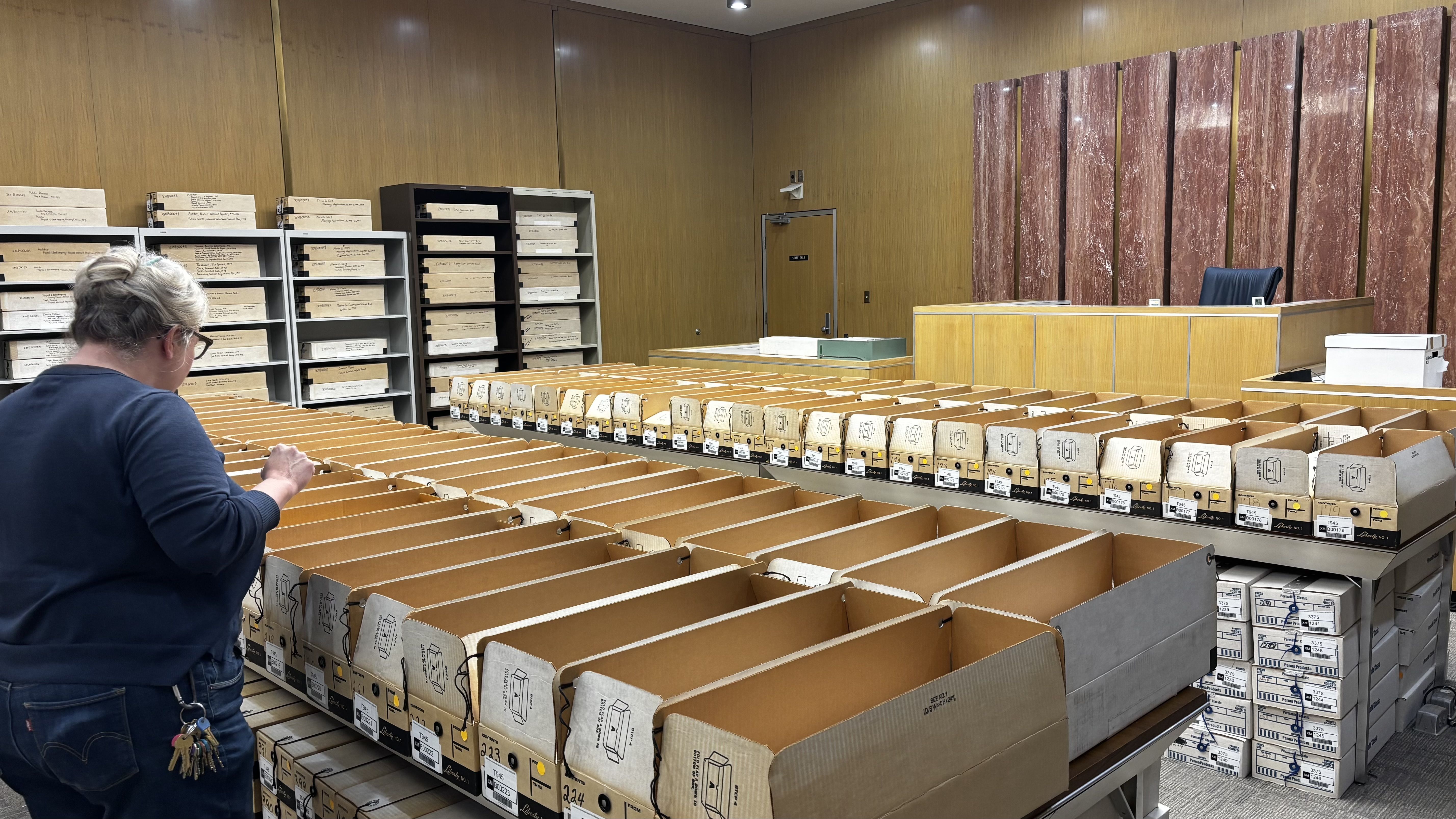 Archive room with a person in a blue shirt inspecting long rows of open tan file boxes on a central table; wood-paneled walls, tall labeled shelves, and a desk with pink marble backdrop.