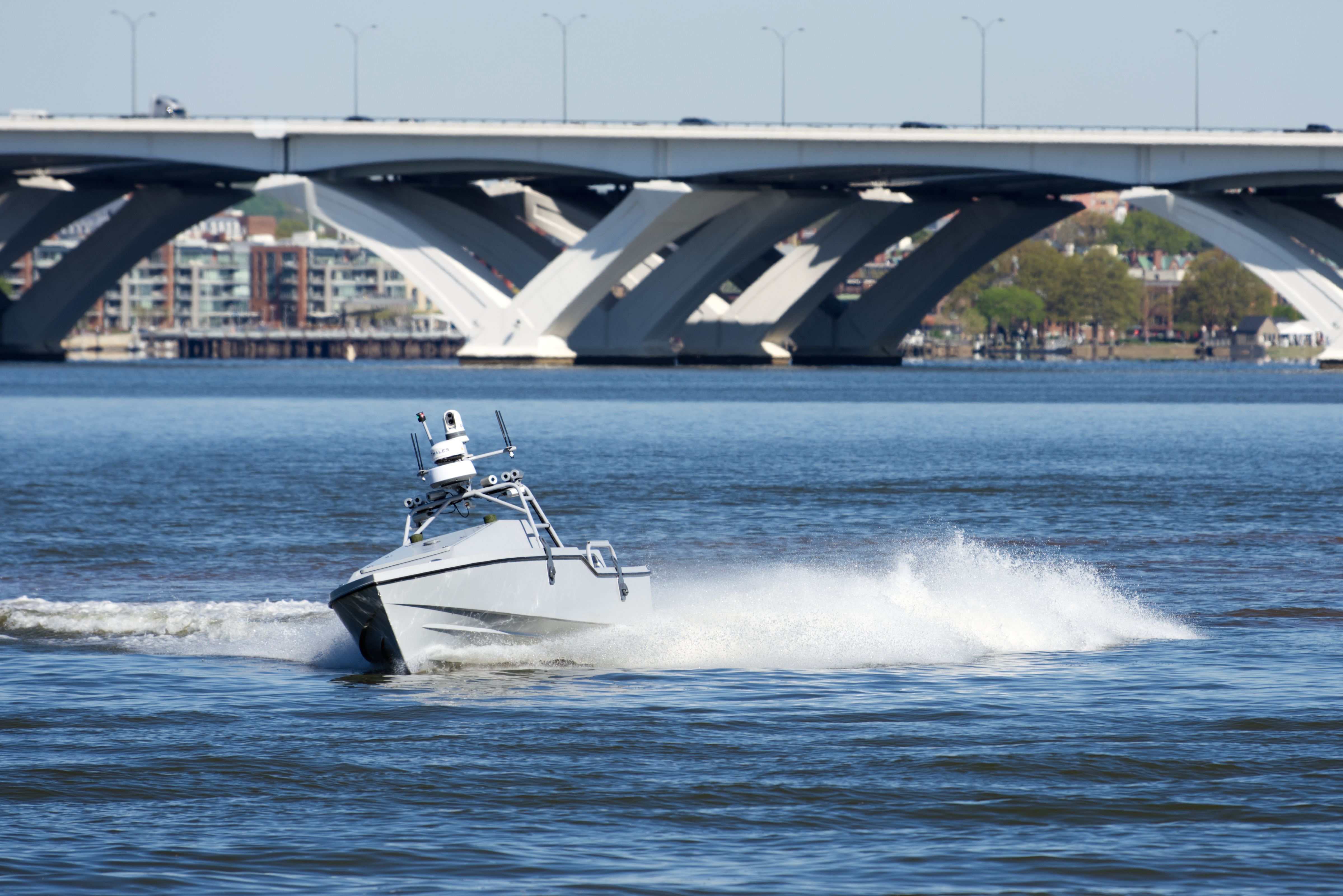 An unmanned boat cuts across the Potomac River. It's kicking up wake. A bridge can be seen in the background.