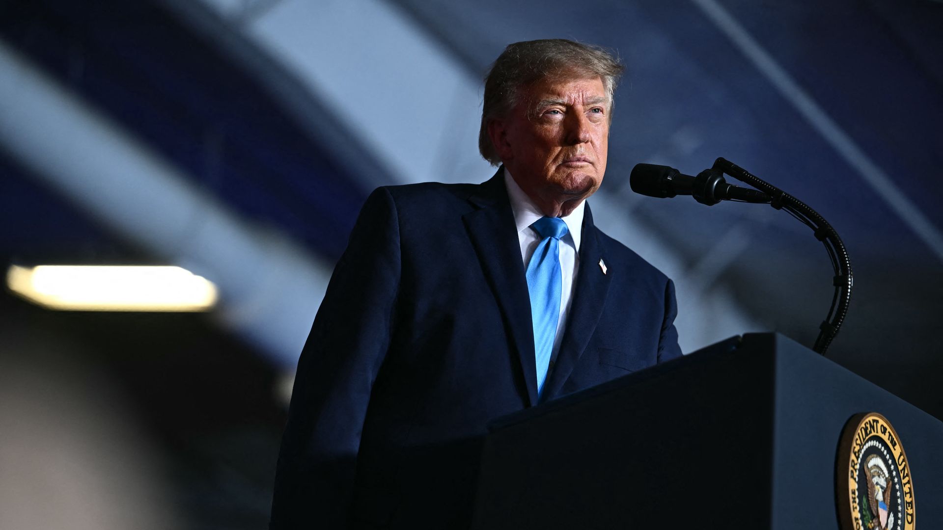 President Trump — wearing a navy blue jacket, a light blue tie, a white collared shirt and an American flag pin — a looks on during a campaign rally while standing behind a podium with a microphone