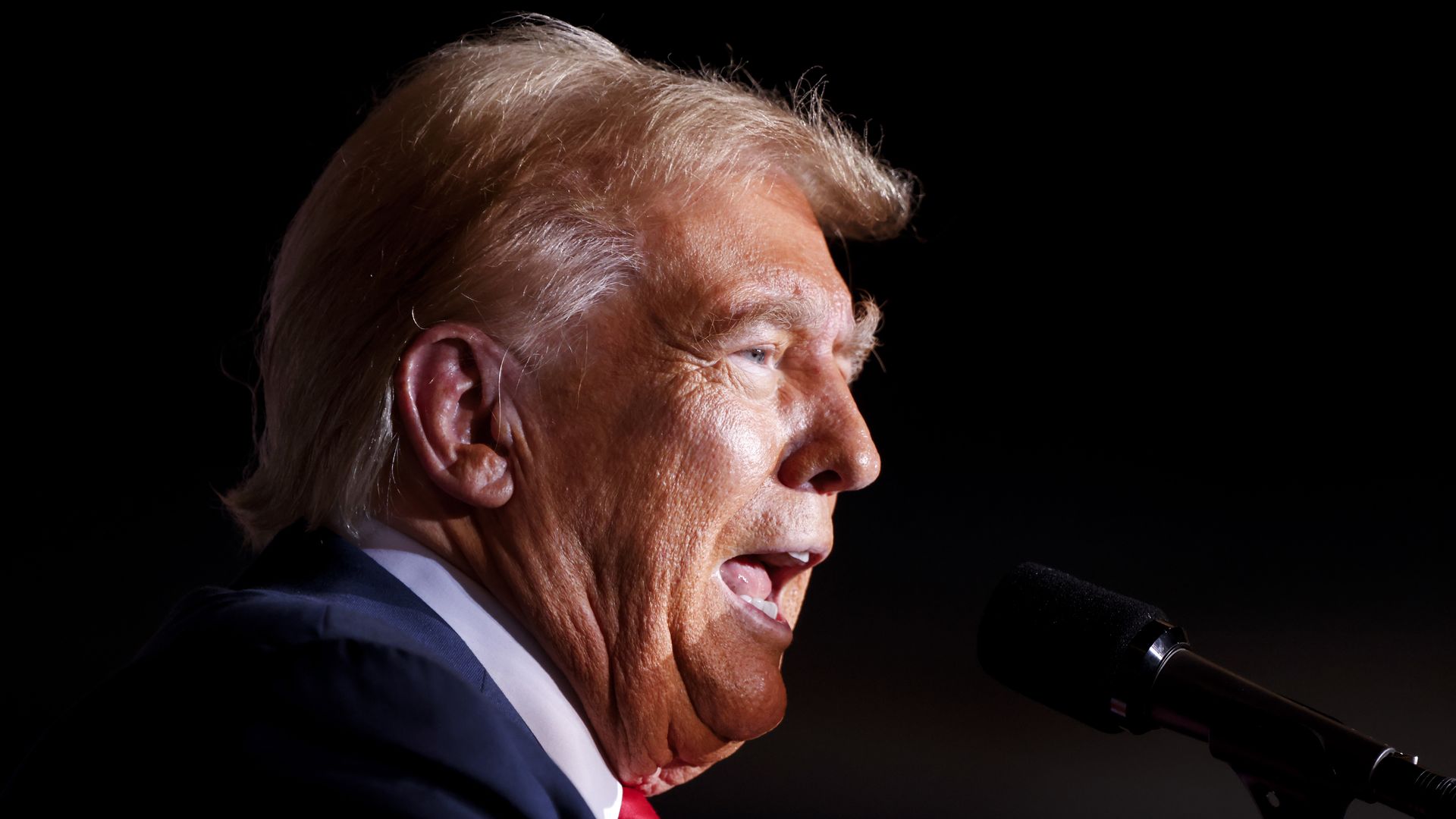 Close-up side profile of a light-haired older man in a dark suit and red tie, speaking into a microphone against a black background.