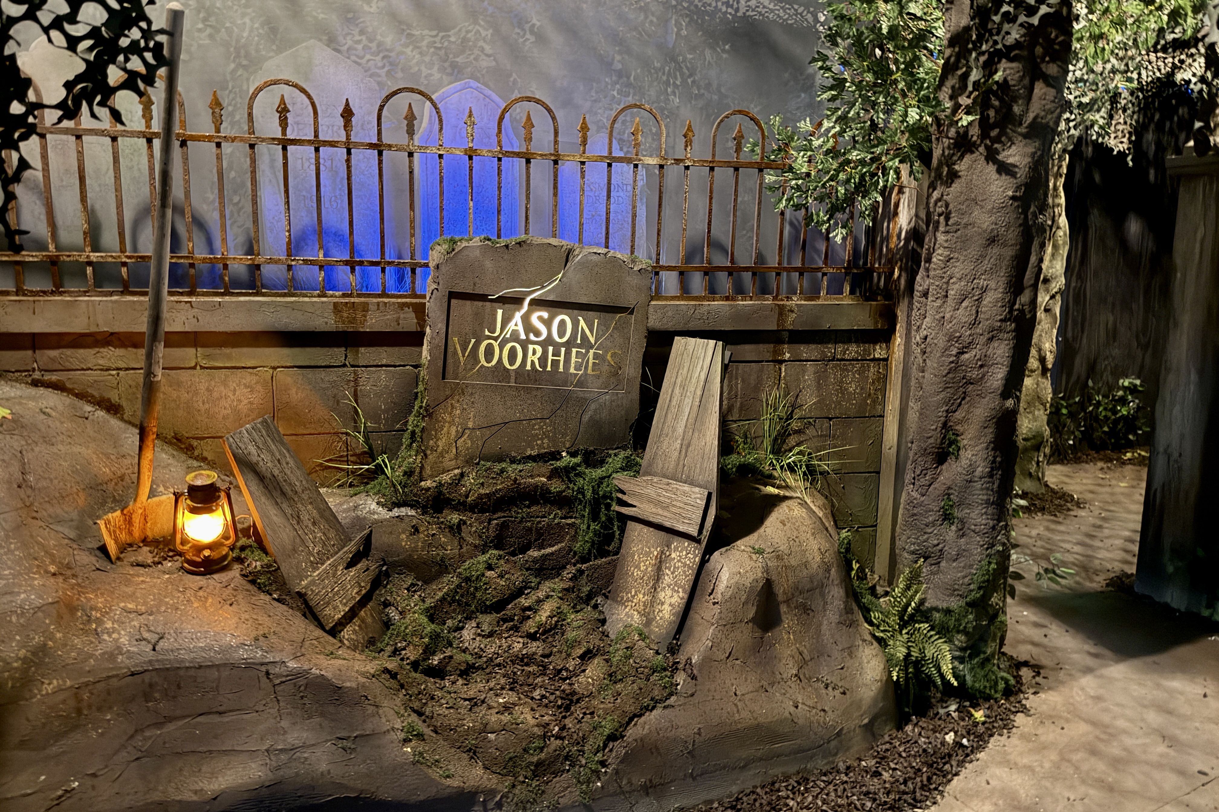 Rusty wrought-iron fence with arched spears blocks a foggy graveyard filled with blue-glowing tombstones; a cracked stone plaque reads "JASON VOORHEES" while a lit lantern and debris sit nearby.