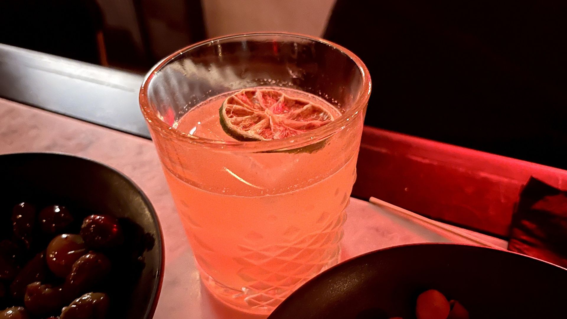 A close-up of a cocktail in a crystal glass with a dried lime on top, sitting next to a bowl of olives and another bowl of nuts. The lighting is a warm, red-ish pink-ish color at Sidecar.