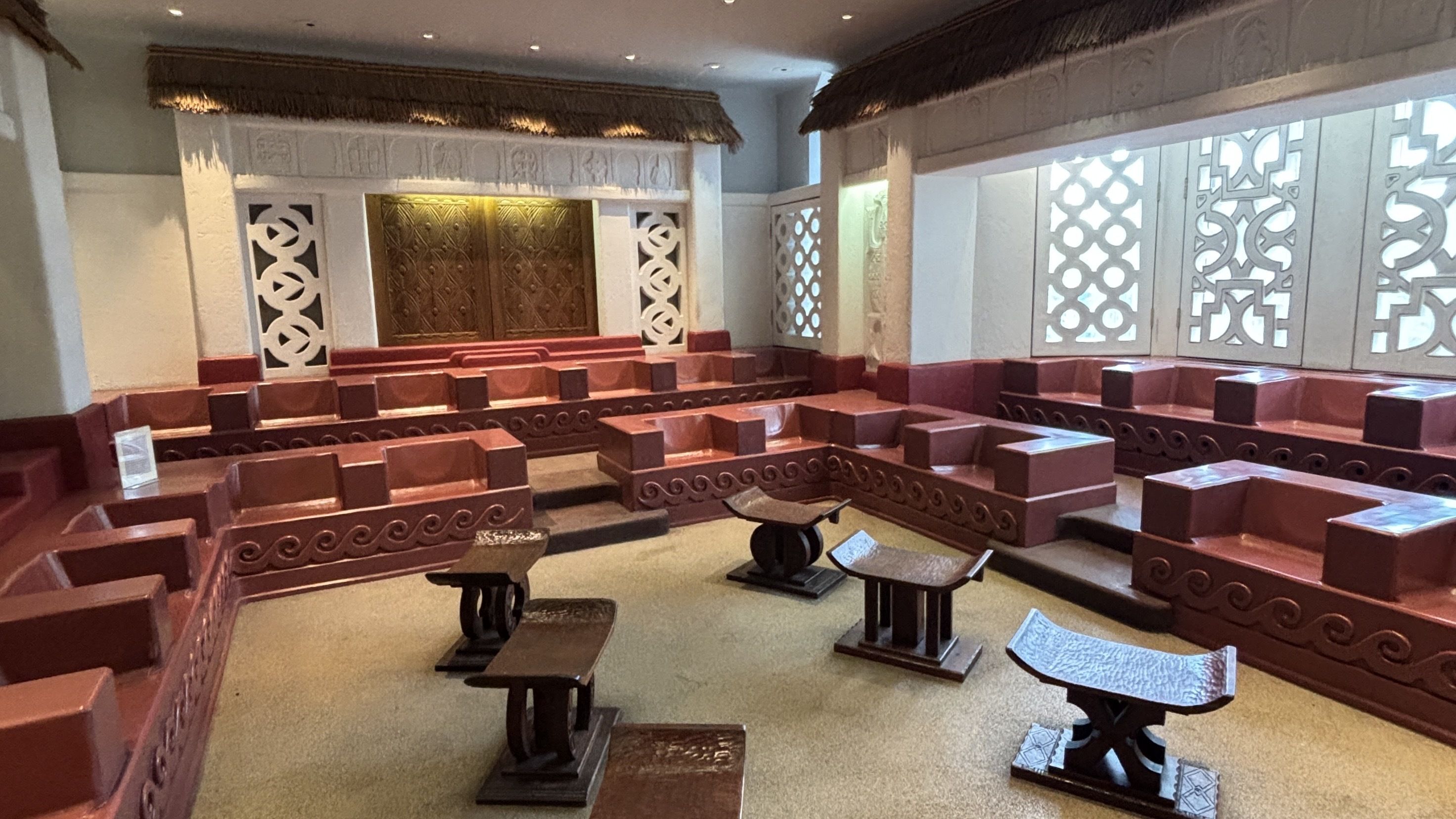 A theater-like hall with red-brown tiered benches surrounding a central stage. White carved walls feature decorative lattice panels, and a thatched roof sweeps over the stage.
