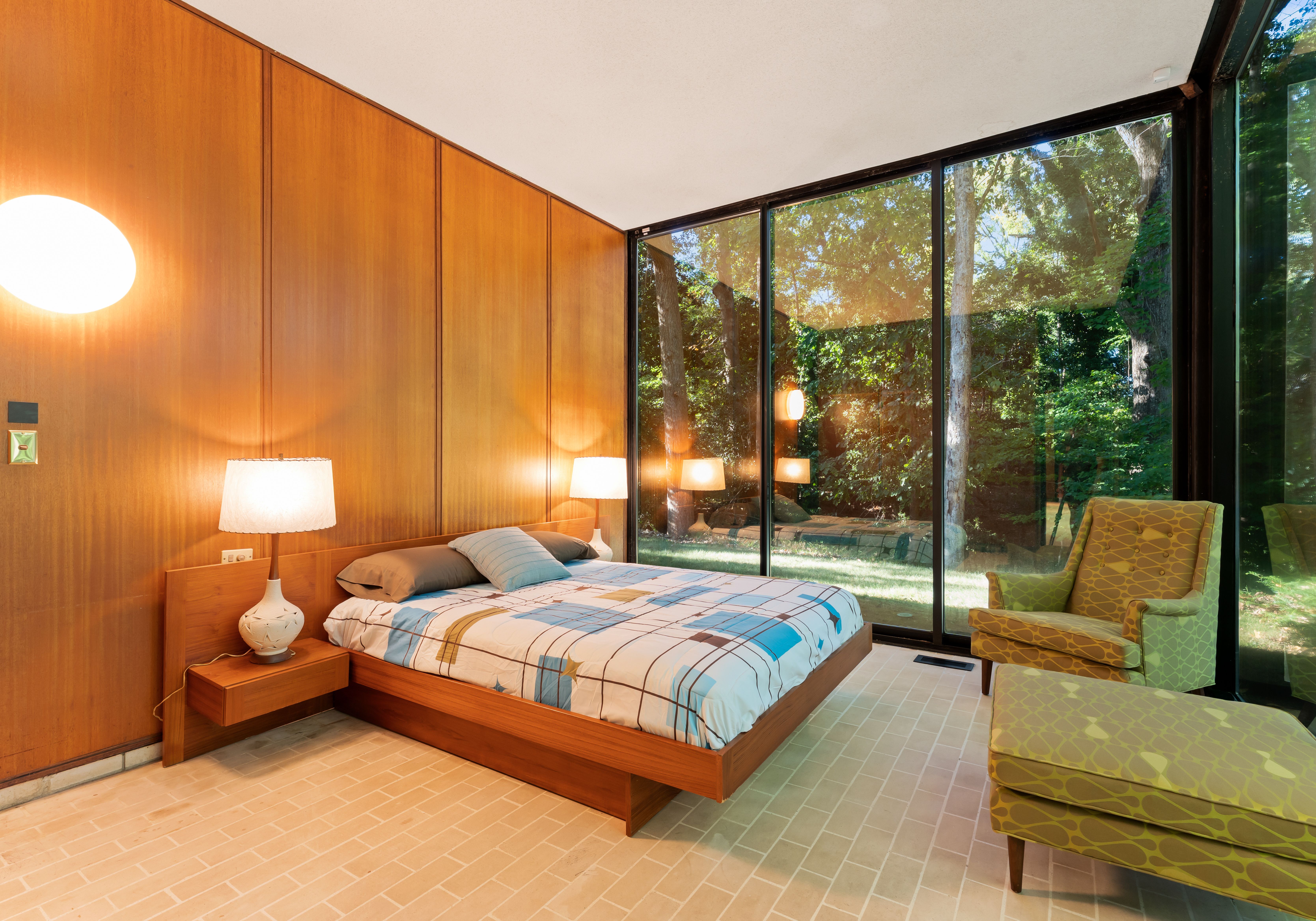 Bedroom with wood-paneled walls, floating wooden bed with patterned blue and beige bedding, two lamps on side tables, large glass windows showing green trees, and green patterned armchair with ottoman.