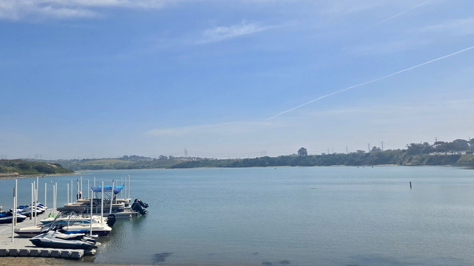 Calm harbor scene with a docked row of boats and jetskis on the left, calm blue water, a distant grassy shoreline, and a bright blue sky with a white contrail streaking across.
