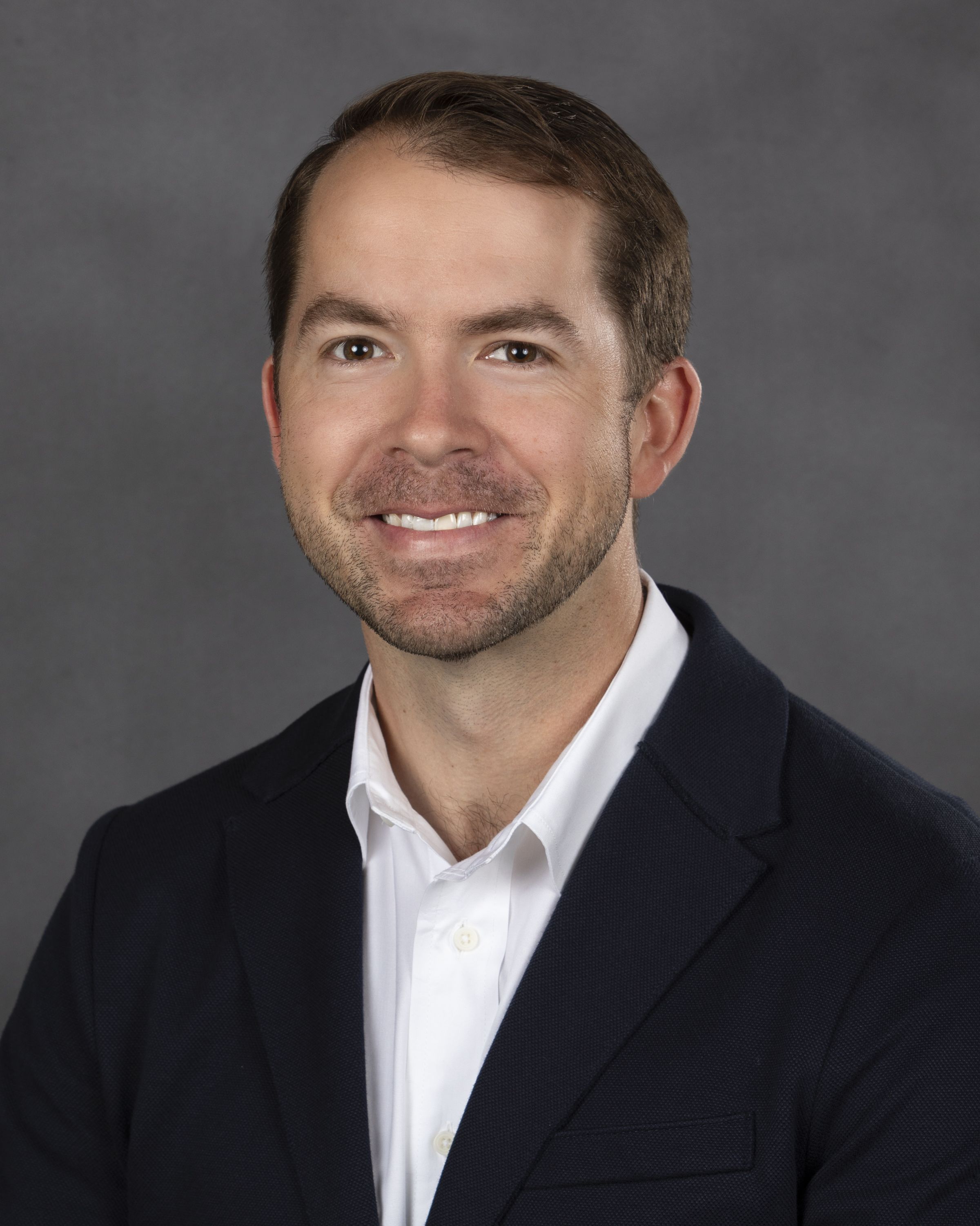 Close-up portrait of a smiling man with short brown hair, wearing a dark blazer and white shirt, against a gray studio backdrop.