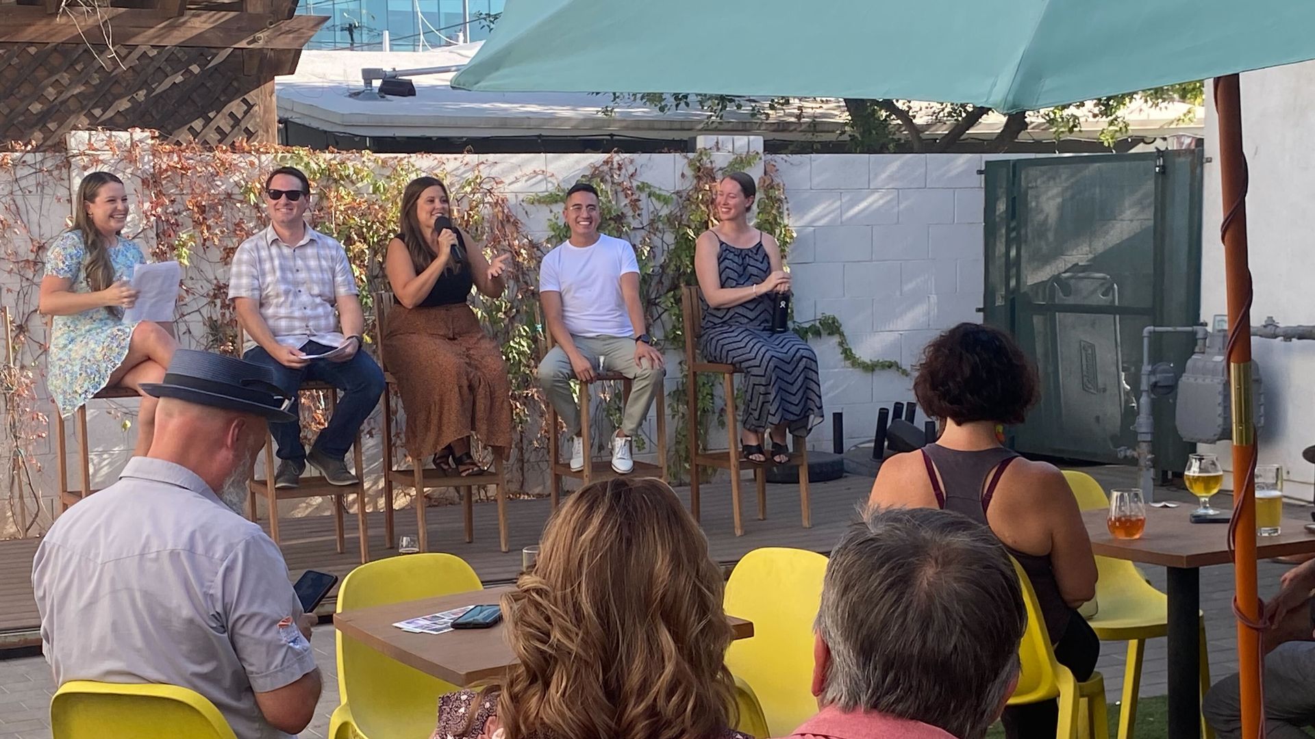 A group of people sitting in yellow metal chairs watches five people sitting in a row on a stage.