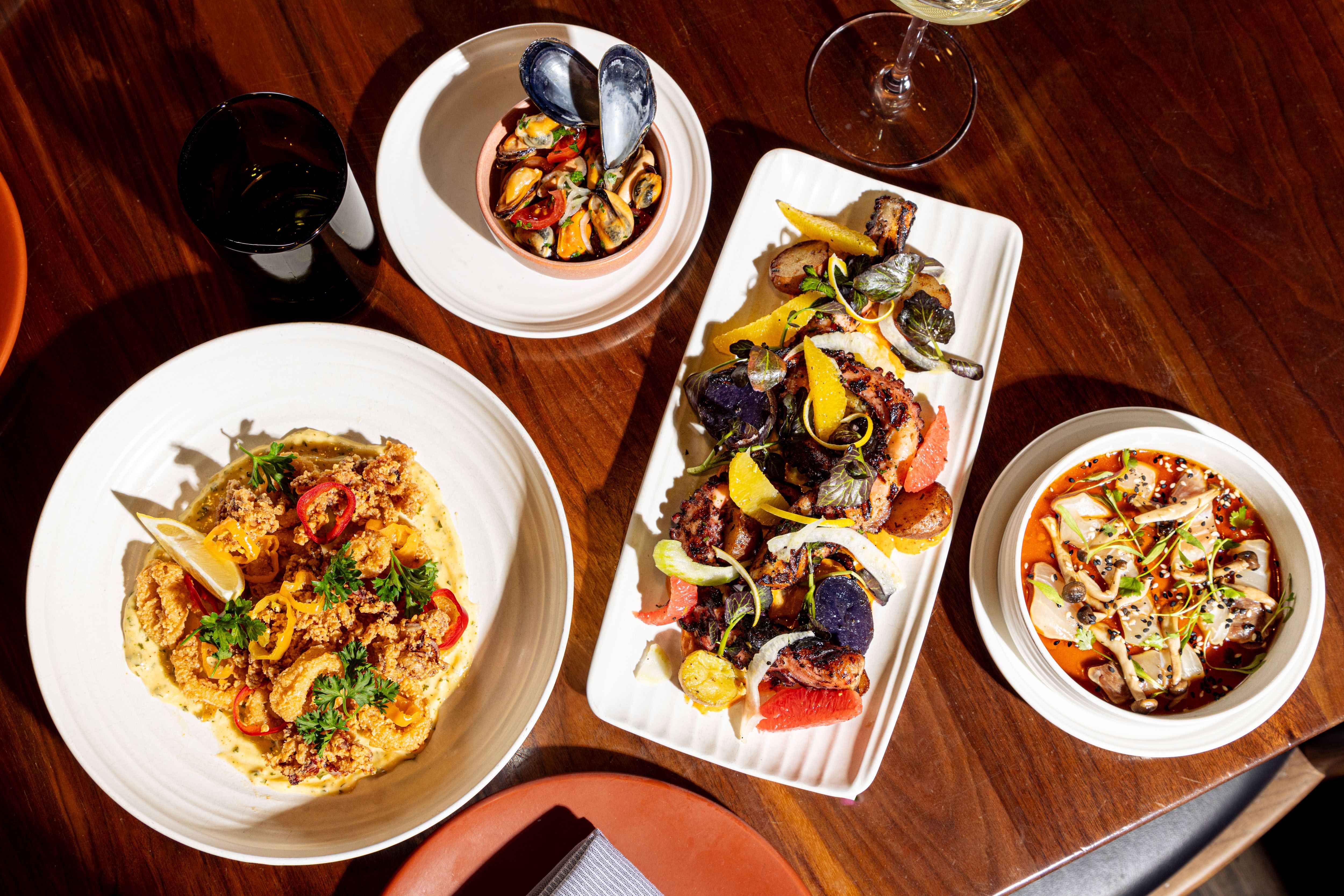 A wooden table with four dishes: fried calamari with lemon and herbs, mussels with tomatoes in a bowl, grilled octopus with citrus and potatoes, and a mushroom dish in red sauce, plus two glasses.