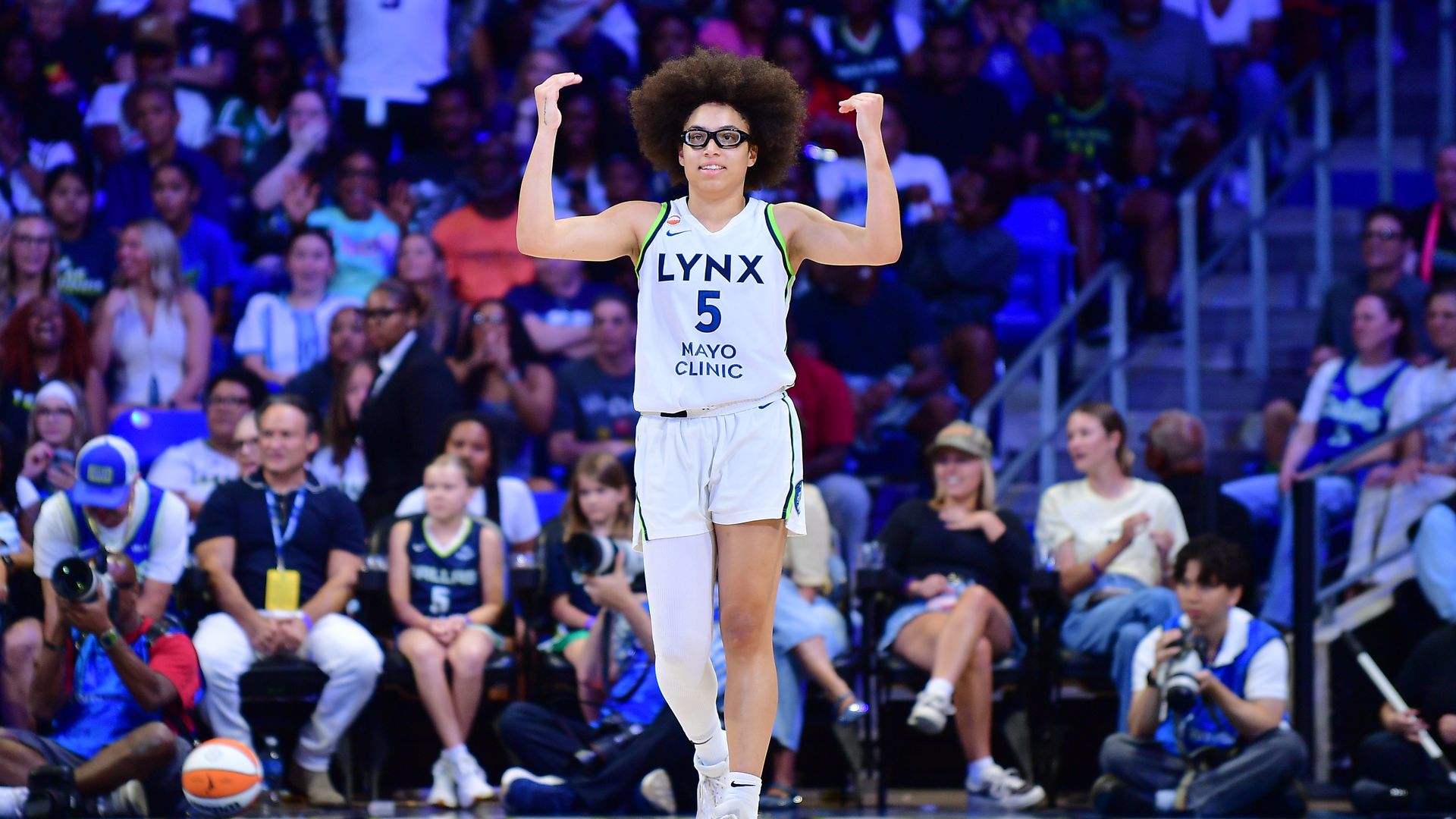Olivia Miles wearing a white Lynx jersey #5, raises arms in victory on a crowded court; blue-lit arena and photographers in background.