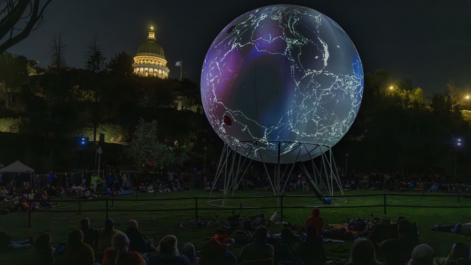 Night in a park: a large illuminated globe on stilts glows blue and purple with white network lines. Crowd sits on blankets while a domed, lit building rises on a hill in the background.