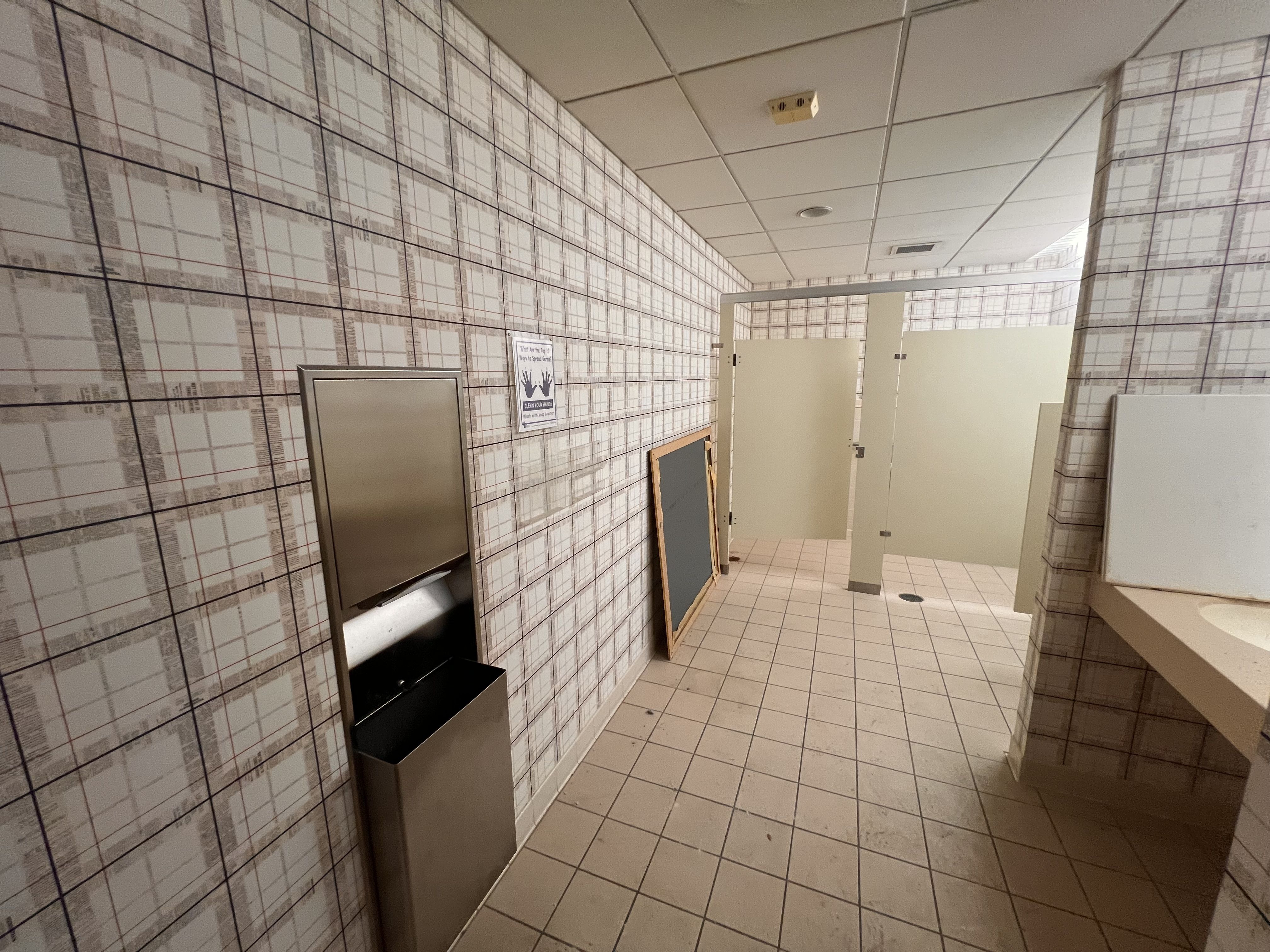 Public restroom with tiled walls and floor, beige stall doors, a row of stalls, a metal hand dryer, and a chalkboard leaning against the wall. A hand-washing poster is visible.