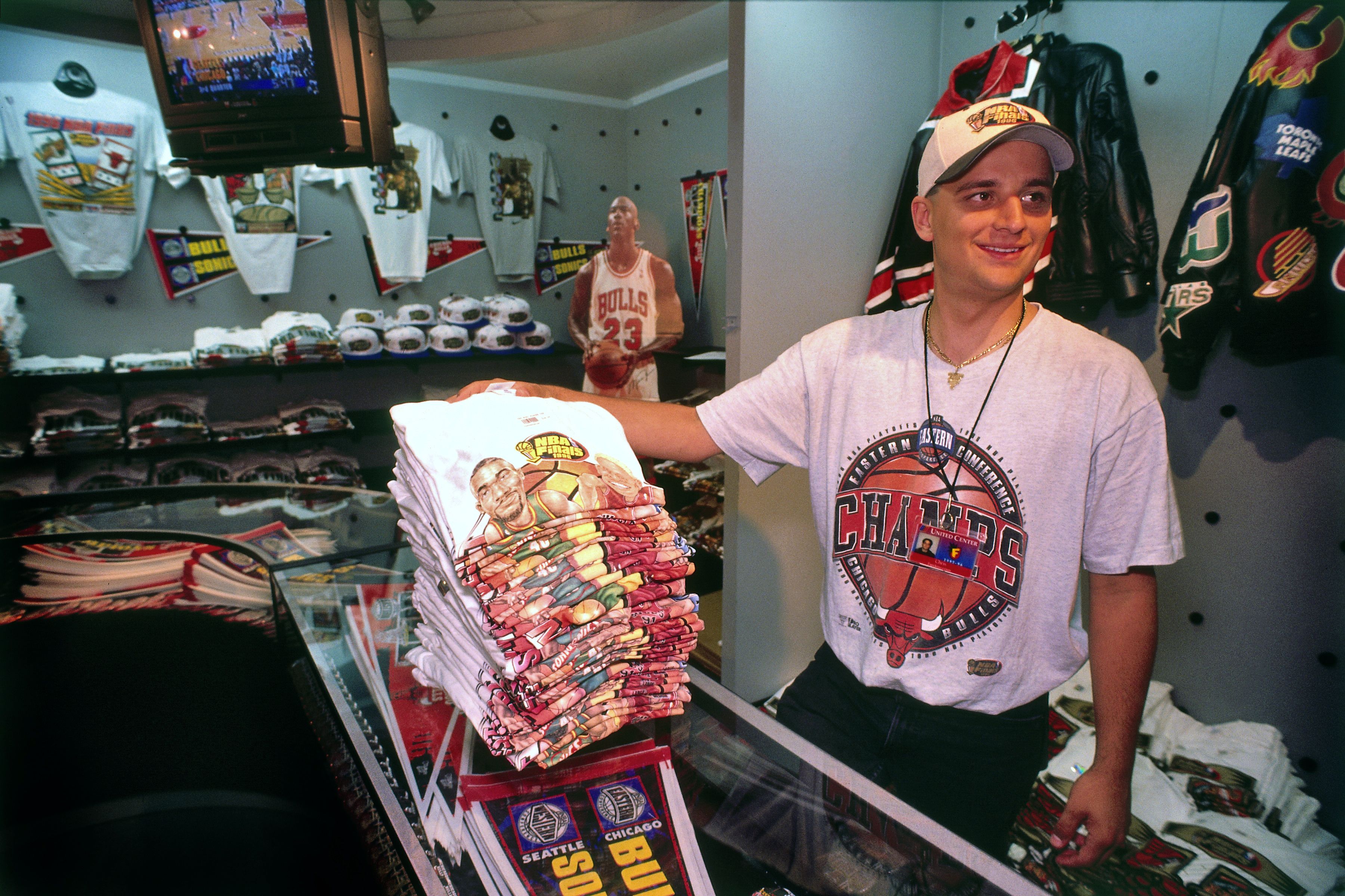 Smiling man in a light gray Bulls T-shirt and cap stands at a glass counter in a retro sports shop, with stacks of tees and a life-size Bulls Michael Jordan cutout in the back.
