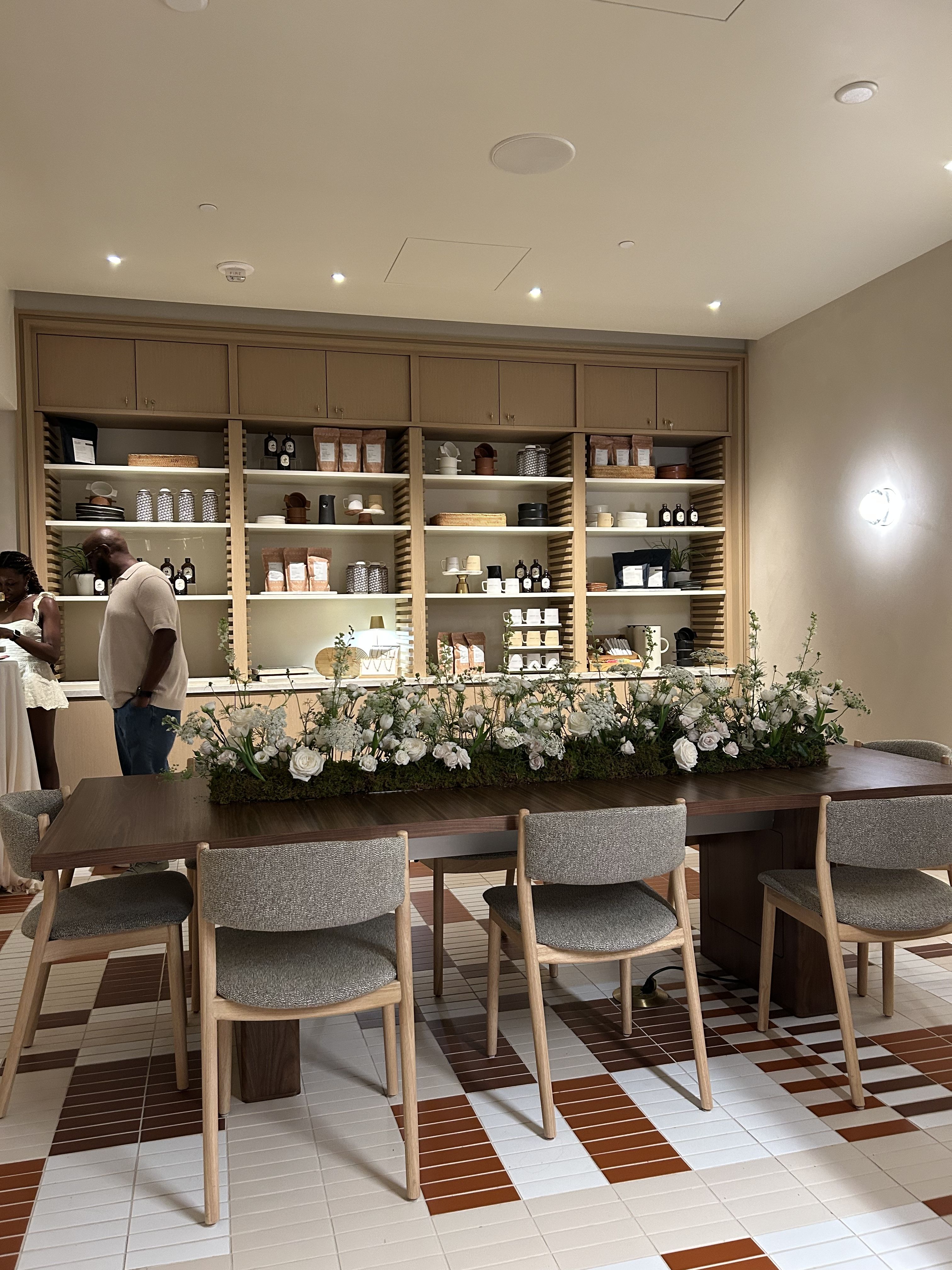Cafe interior with a long wooden table, central floral arrangement, grey chairs, and back shelves of coffee bags and cups; beige walls and a checkered white-brown tiled floor under warm lights.