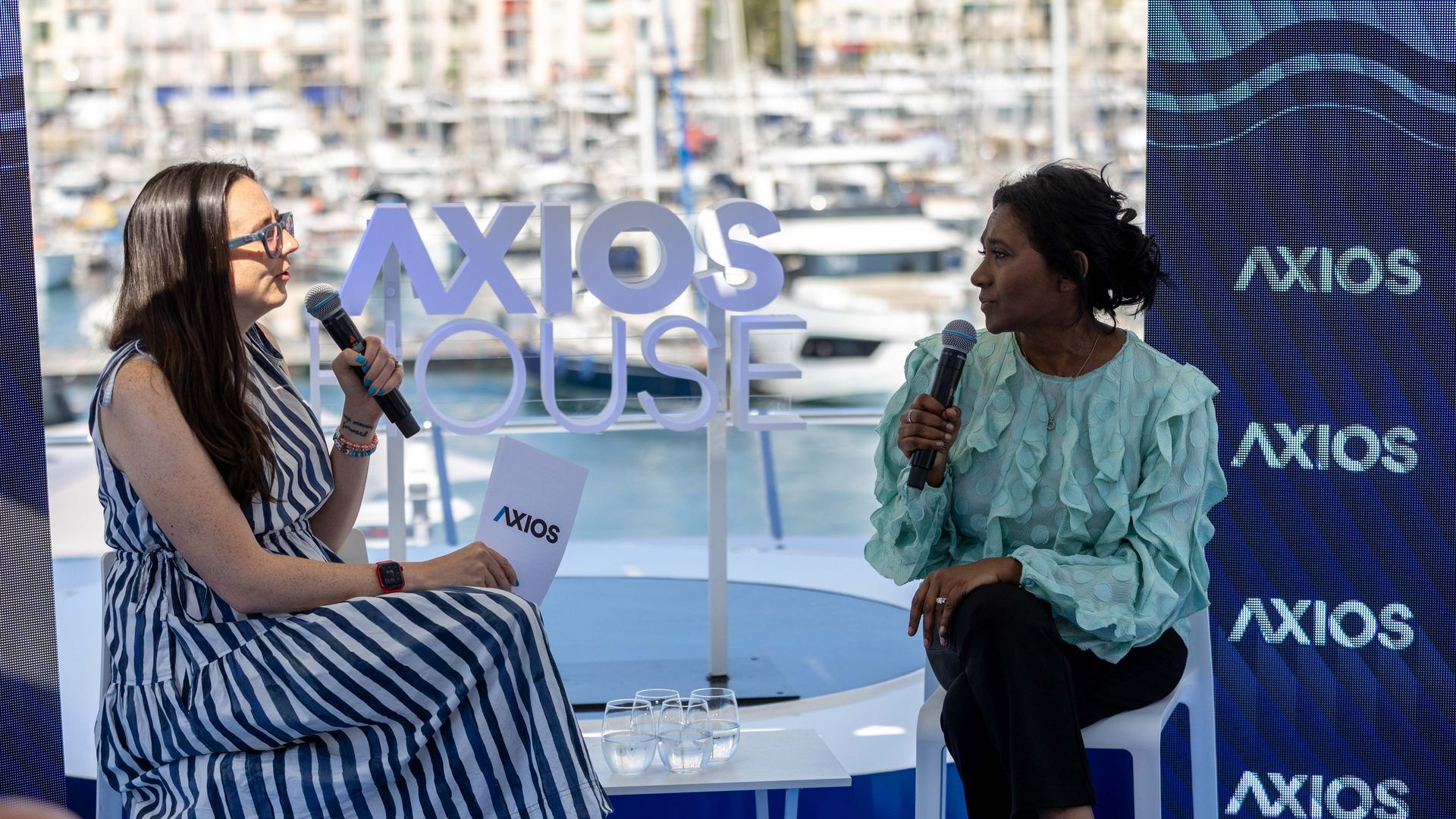 Two women on a panel by a marina, each holding a microphone. Left wears a striped blue-and-white dress and glasses; right wears a mint green blouse. Large AXIOS signs and boats in background.