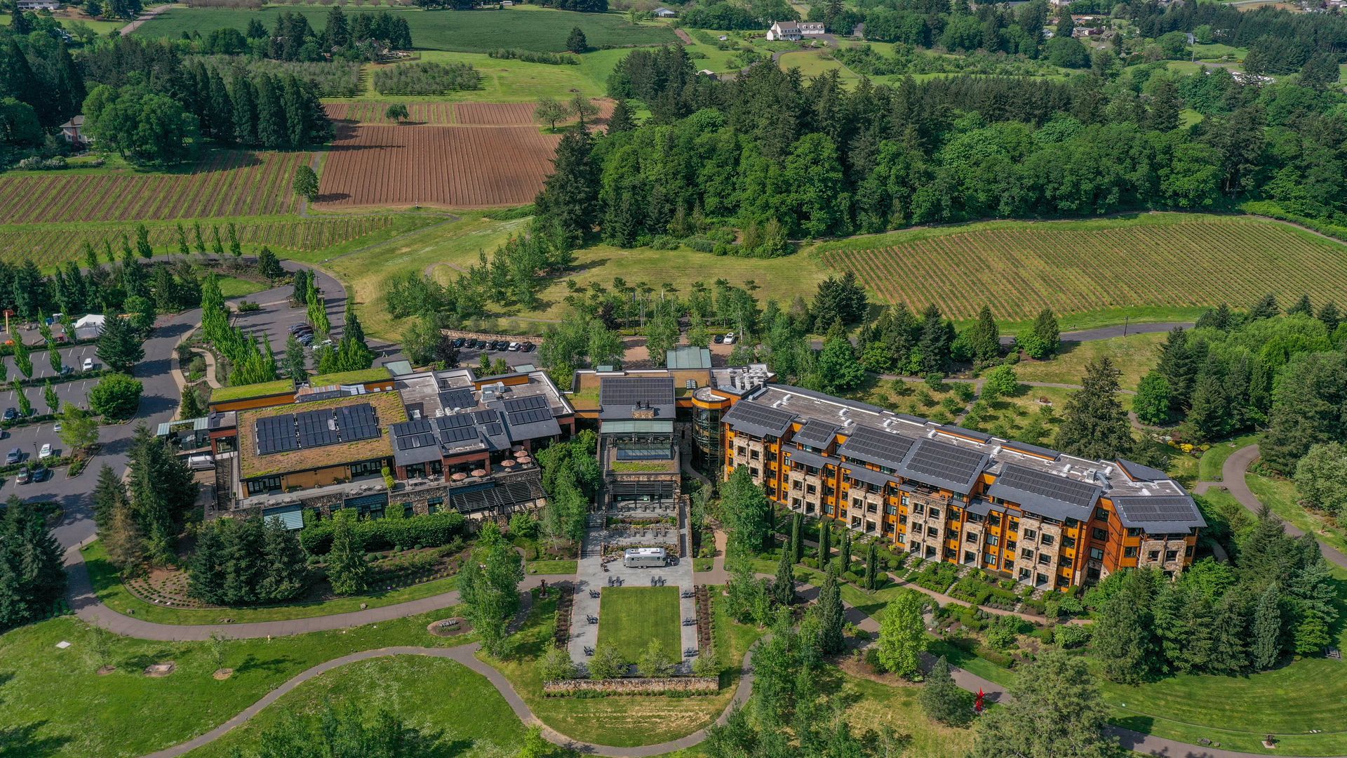 Aerial view of a large building with solar panels on the roof surrounded by lush green lawns, trees, and farmland with vineyards and rural houses in the background.
