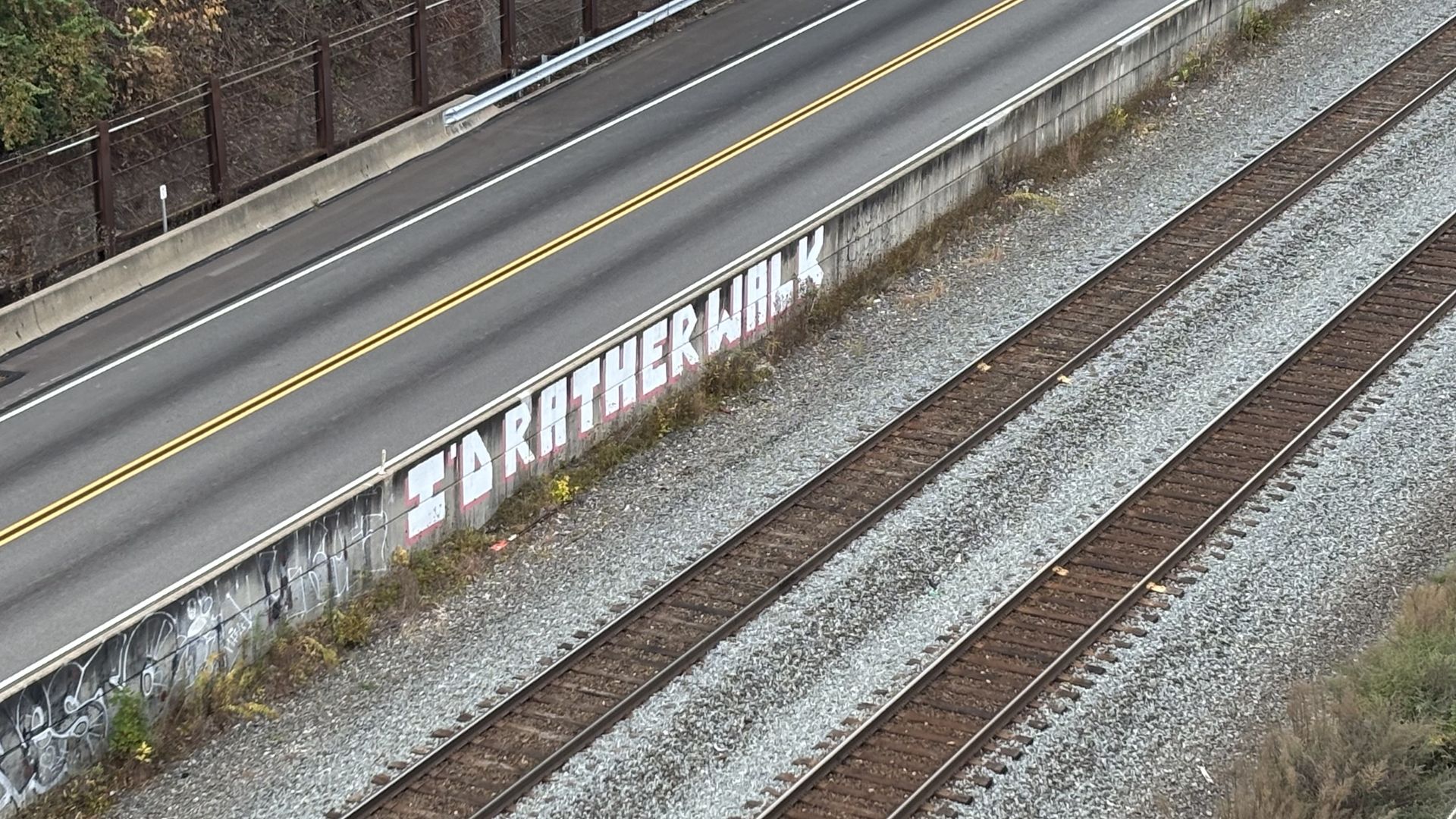 Empty road and railroad tracks separated by a wall with graffiti saying "I'D RATHER WALK" in large white letters outlined in red.