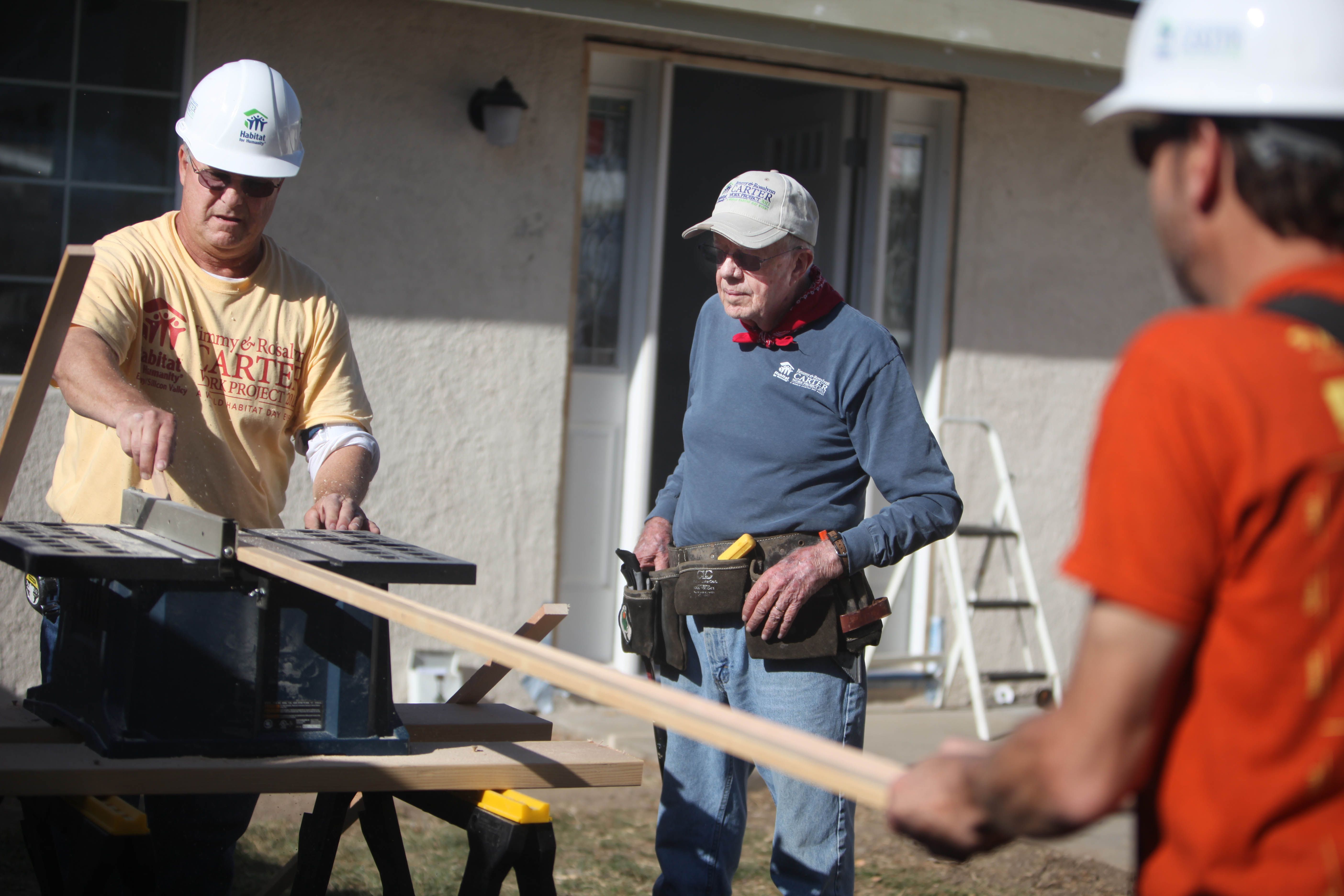 Jimmy Carter in a blue sweatshirt and gray hat, wearing a tool belt around his waist, assisting Habitat workers