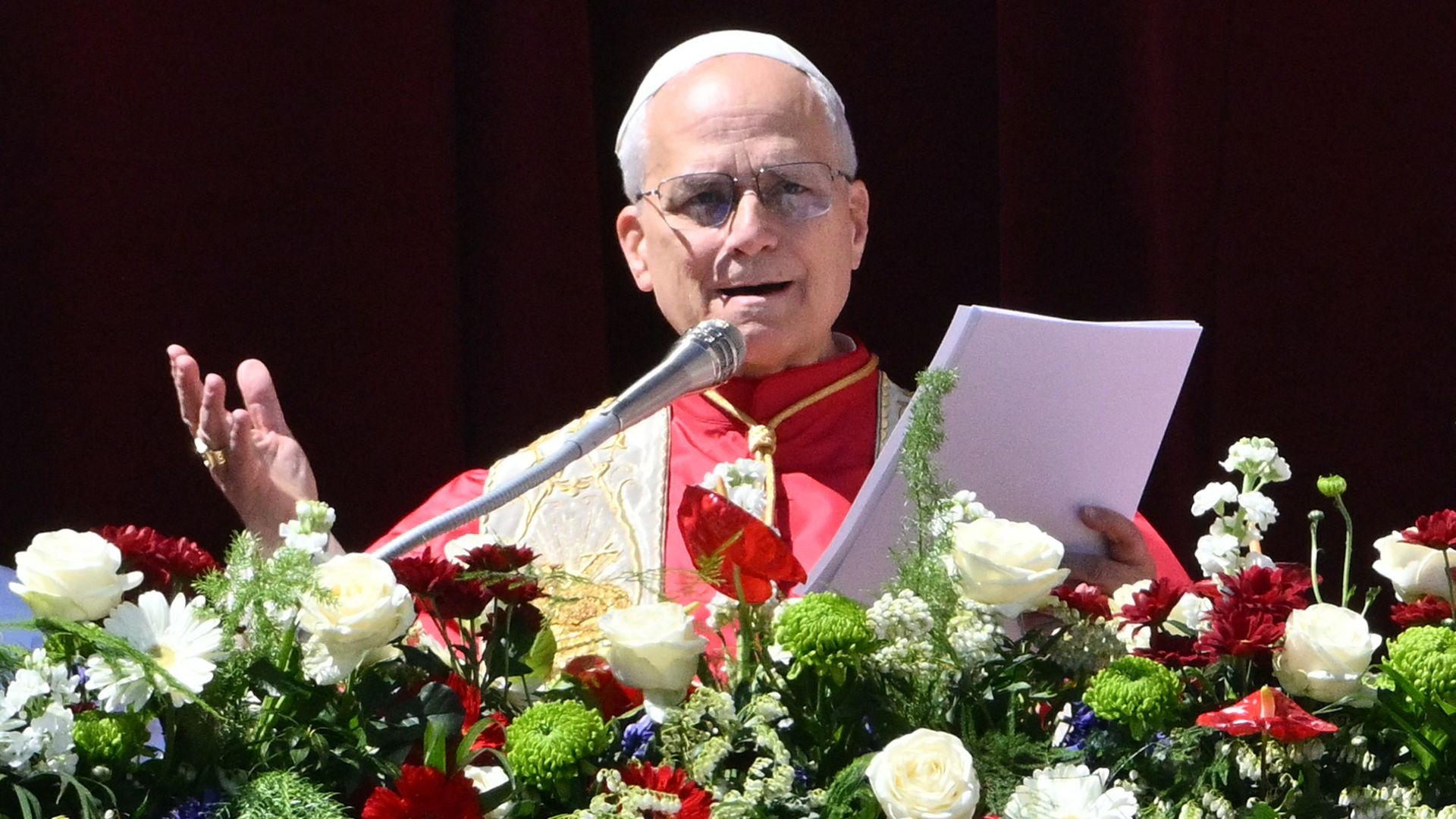 Pope Leo in red robes and a white skullcap speaks into a microphone, holding papers, with a vibrant arrangement of white, red, and green flowers in front.