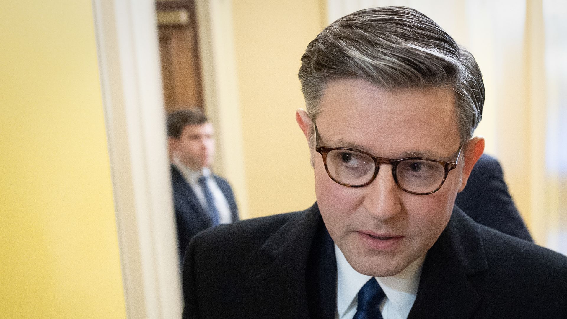 Man with salt-and-pepper hair and brown glasses in a dark suit and blue striped tie, looking downward in a yellow hallway with a blurred person in the background.
