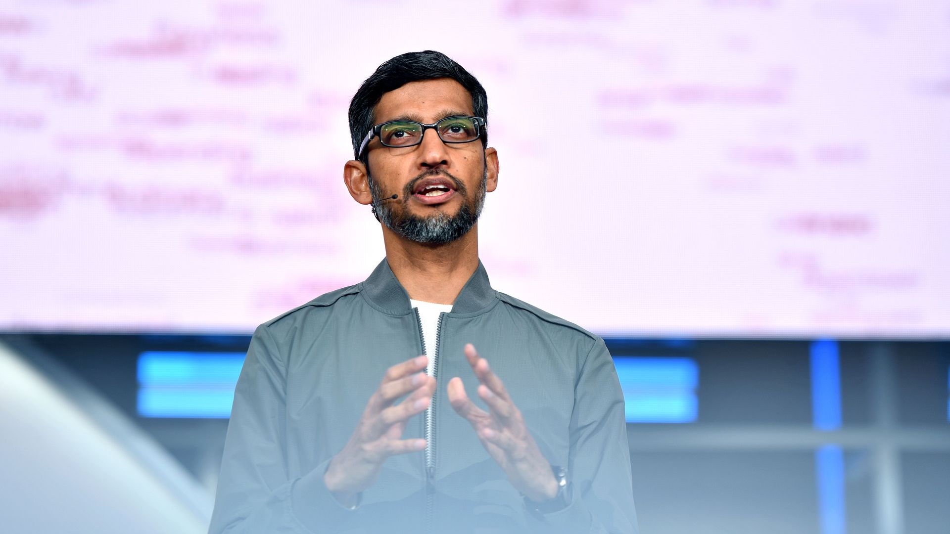 In this image, Sundar Pichai stands on stage and speaks to an audience while wearing a microphone and glasses