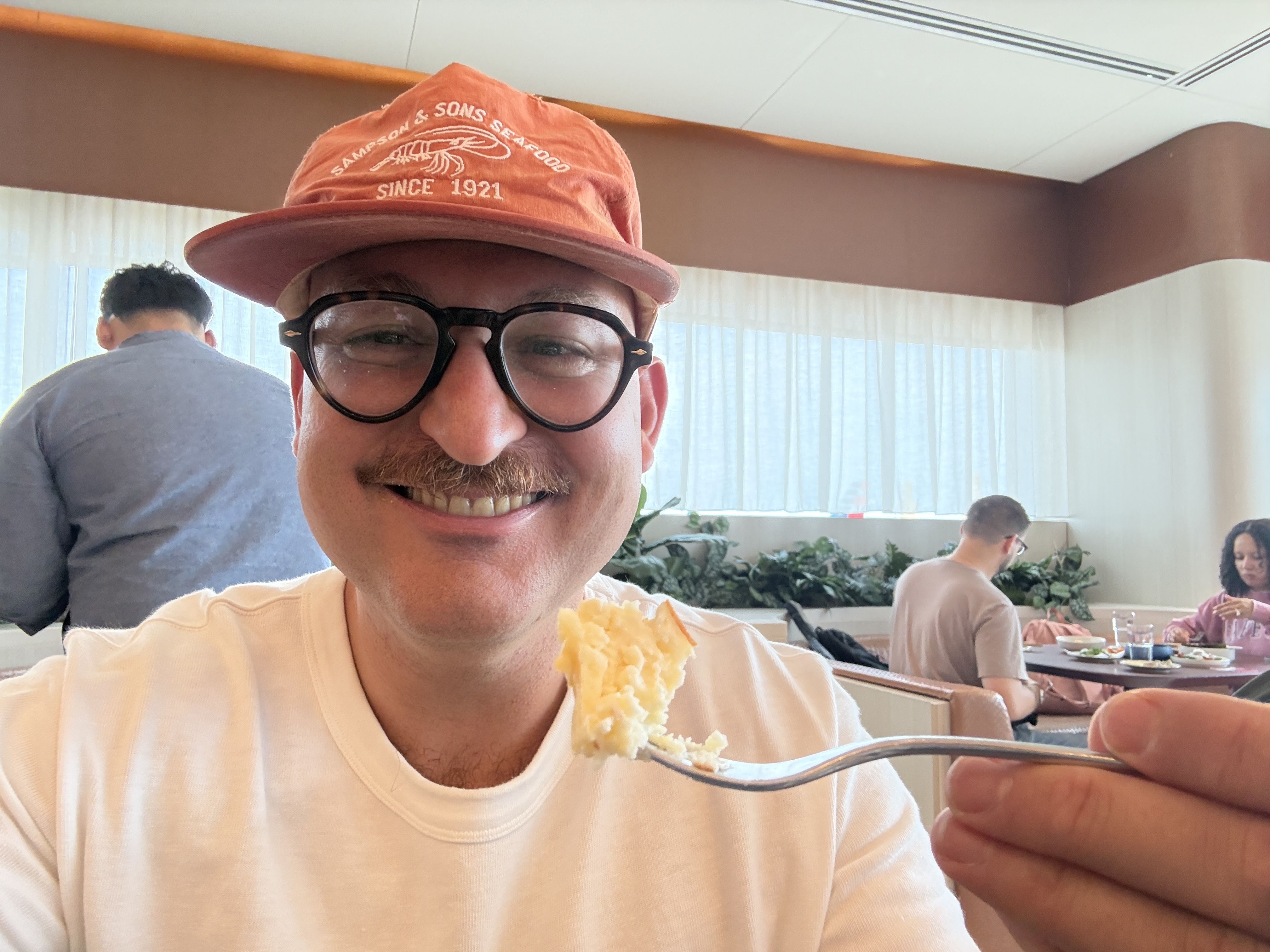 Close-up of a person wearing an orange seafood-logo hat and black round glasses, smiling with a bite of pale food on a fork; diners and plants peek from a bright restaurant behind.